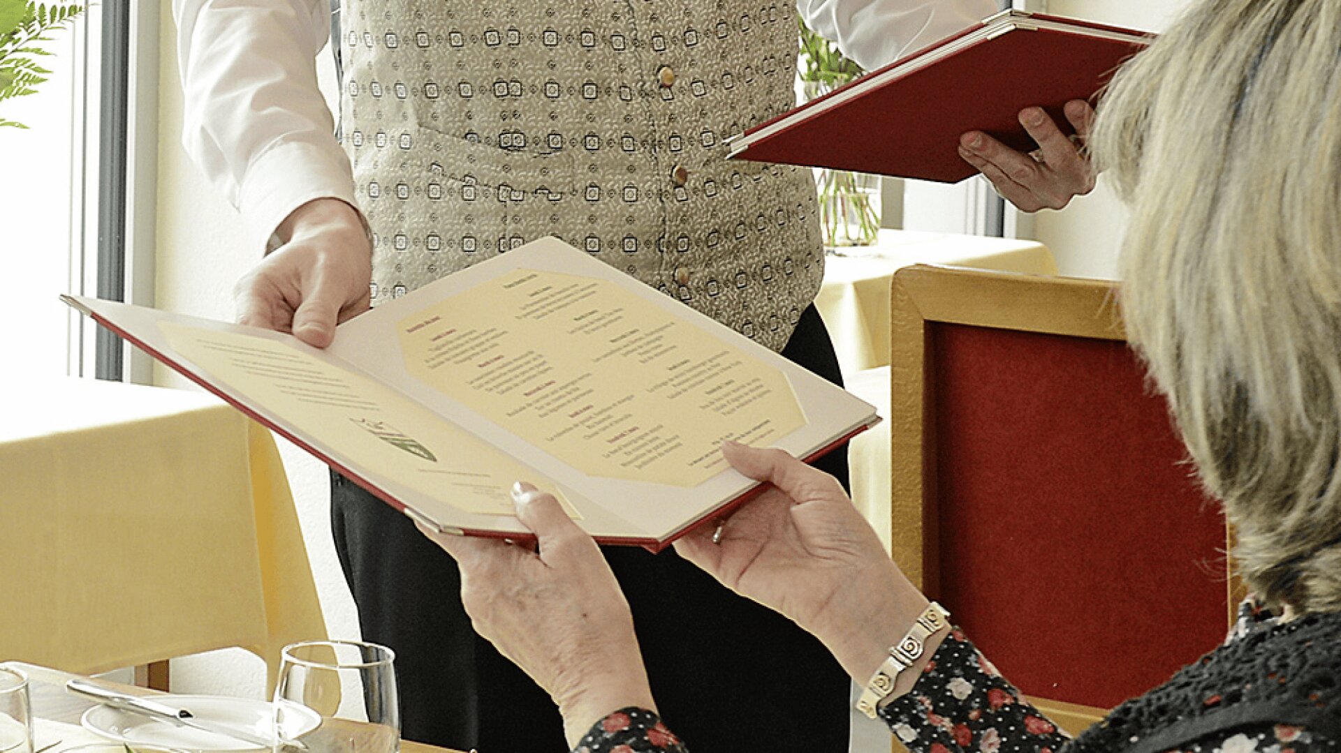 Une personne assise à une table dans un restaurant tient un menu ouvert tandis qu'un serveur vêtu d'un gilet à motifs tient un autre menu, prêt à prendre la commande.