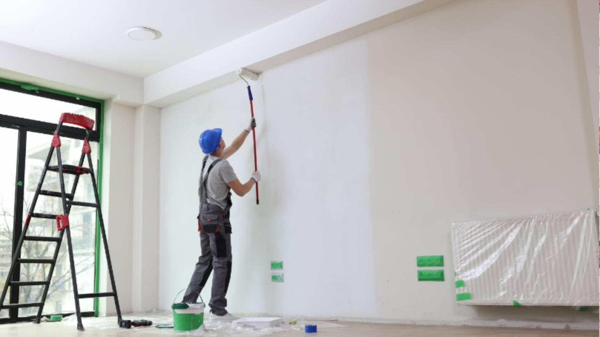 Une personne portant un casque bleu utilise un rouleau pour peindre un mur blanc dans une pièce. Un seau de peinture vert, une échelle et un radiateur couvert se trouvent à proximité. Certaines prises de courant et certains bords sont protégés par du ruban adhésif.