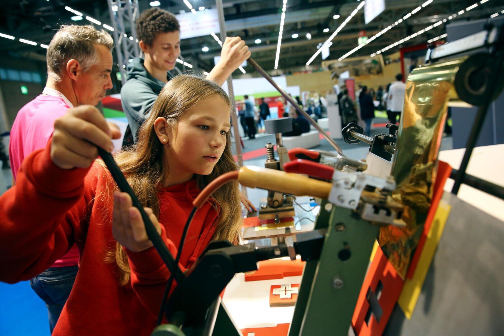 Une jeune fille fait fonctionner une machine dans une exposition scientifique ou technologique interactive, tandis que deux personnes se tiennent à proximité pour observer et participer. Il semble s'agir d'un événement ou d'une exposition en intérieur avec plusieurs stands.