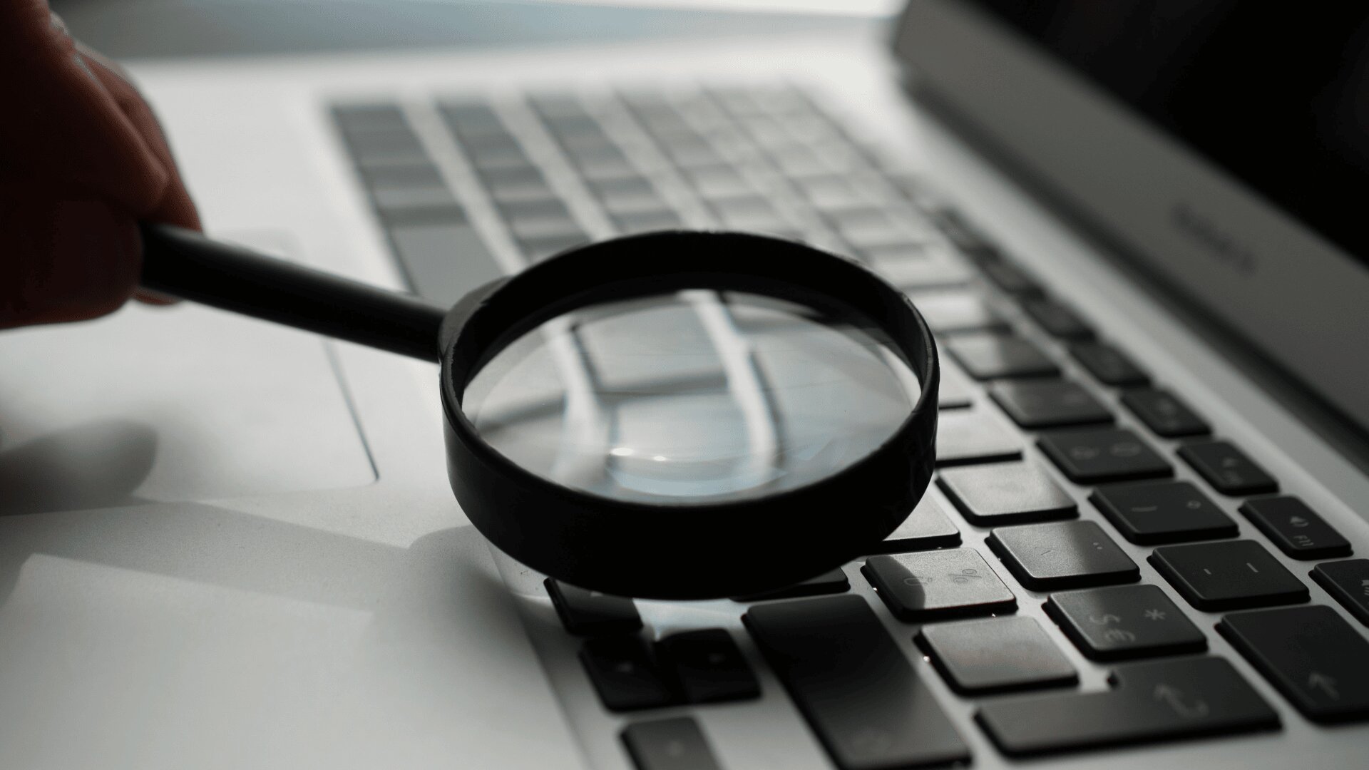 Une main tient une loupe au-dessus du clavier d'un ordinateur portable, se concentrant sur les touches et le pavé tactile, symbolisant la recherche ou l'investigation sur un ordinateur.
