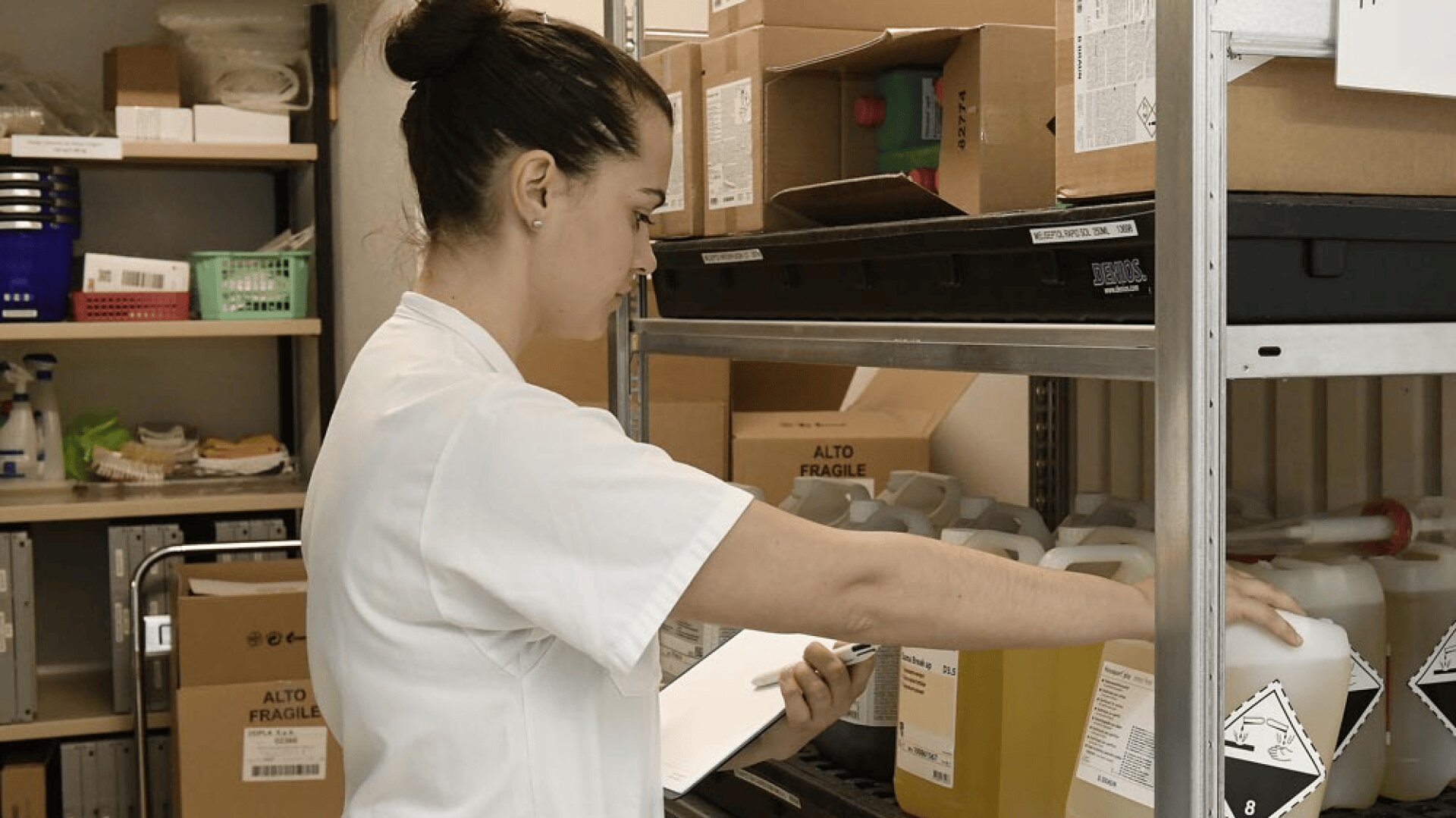 Une femme vêtue d'une blouse blanche vérifie l'inventaire sur un presse-papiers à côté d'étagères remplies de boîtes et de grands conteneurs en plastique à l'intérieur d'une salle d'entreposage.