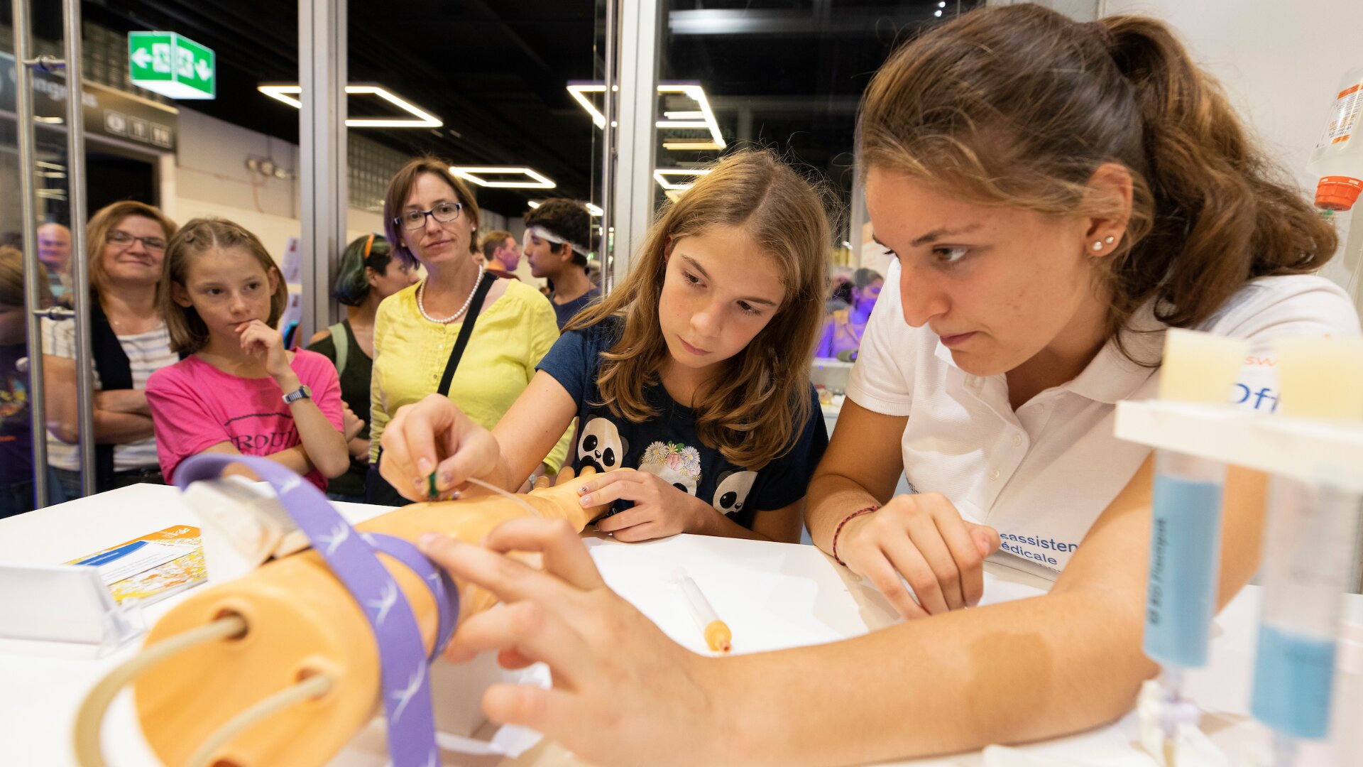Deux jeunes filles pratiquent des actes médicaux sur le bras d'un mannequin pendant qu'une femme en polo blanc les guide. Plusieurs personnes les observent attentivement à l'arrière-plan, dans une pièce très éclairée.