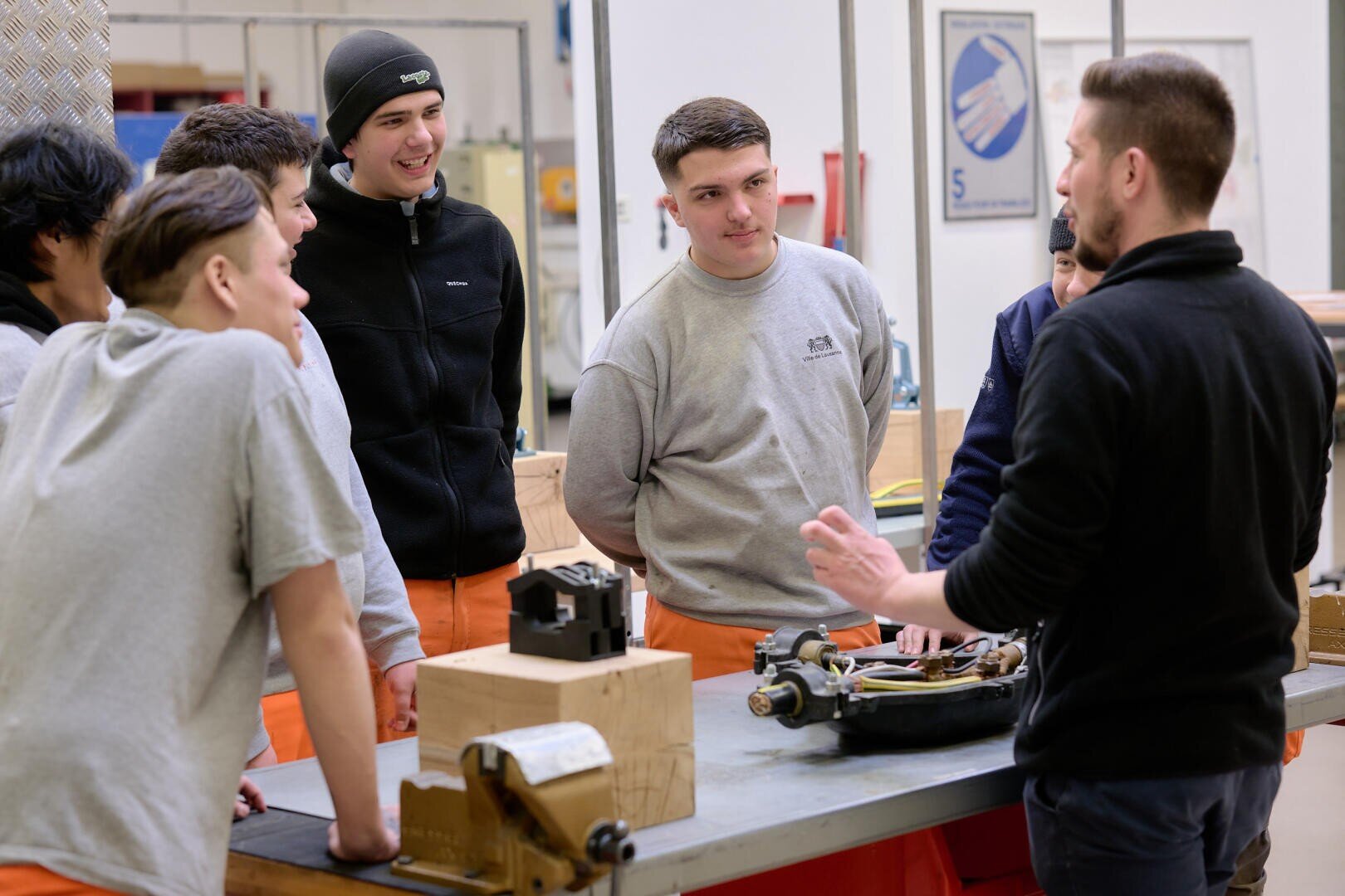 Un groupe de jeunes hommes écoute attentivement un instructeur expliquer un dispositif mécanique dans un atelier, avec des outils et des équipements visibles sur la table.