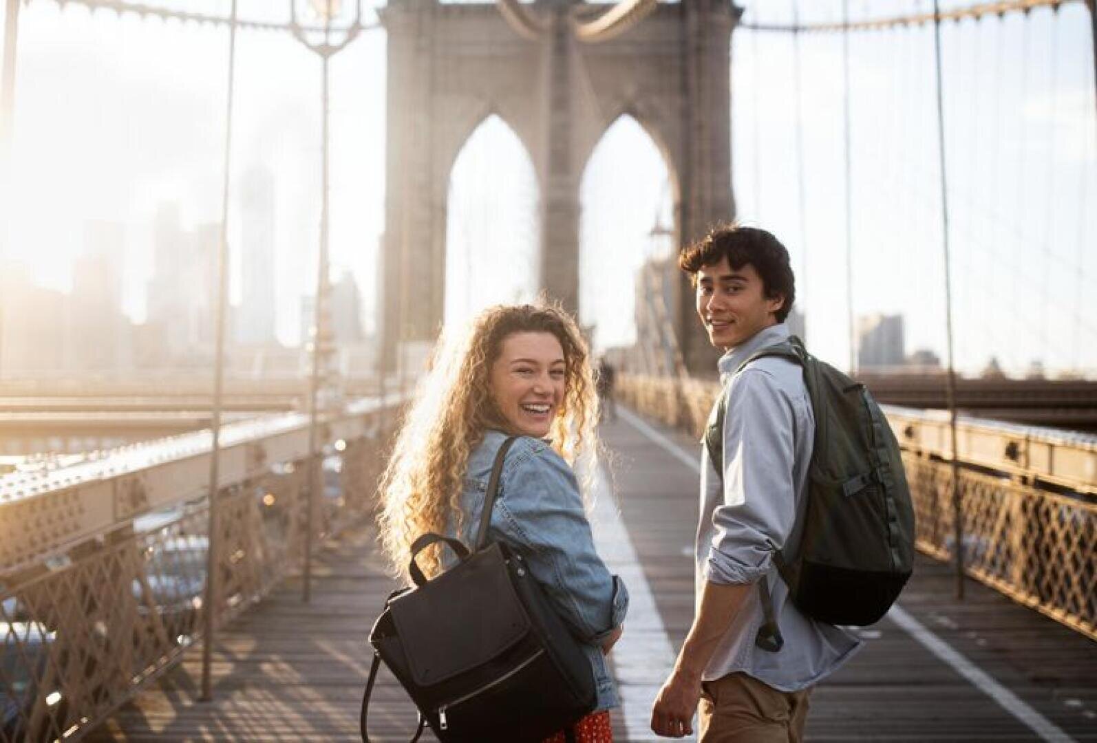 Une jeune femme et un homme portant un sac à dos sourient en marchant ensemble sur le pont de Brooklyn, à New York, au lever ou au coucher du soleil. La ligne d'horizon de la ville est visible en arrière-plan.