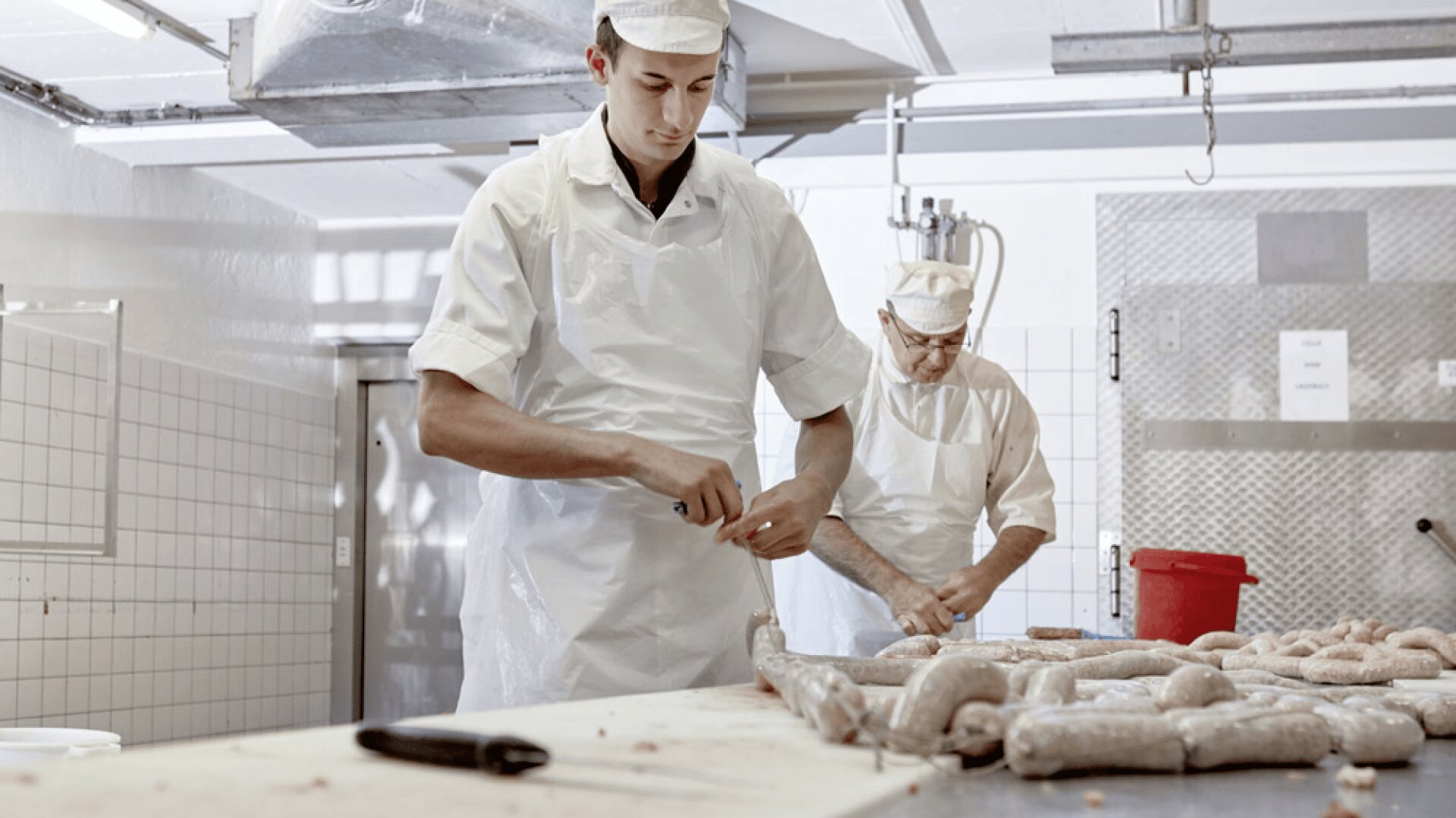Deux bouchers portant des tabliers et des chapeaux blancs préparent des saucisses dans une cuisine propre et carrelée. L'un d'eux tord des saucisses au premier plan, tandis que l'autre travaille à l'arrière-plan près d'un seau rouge.