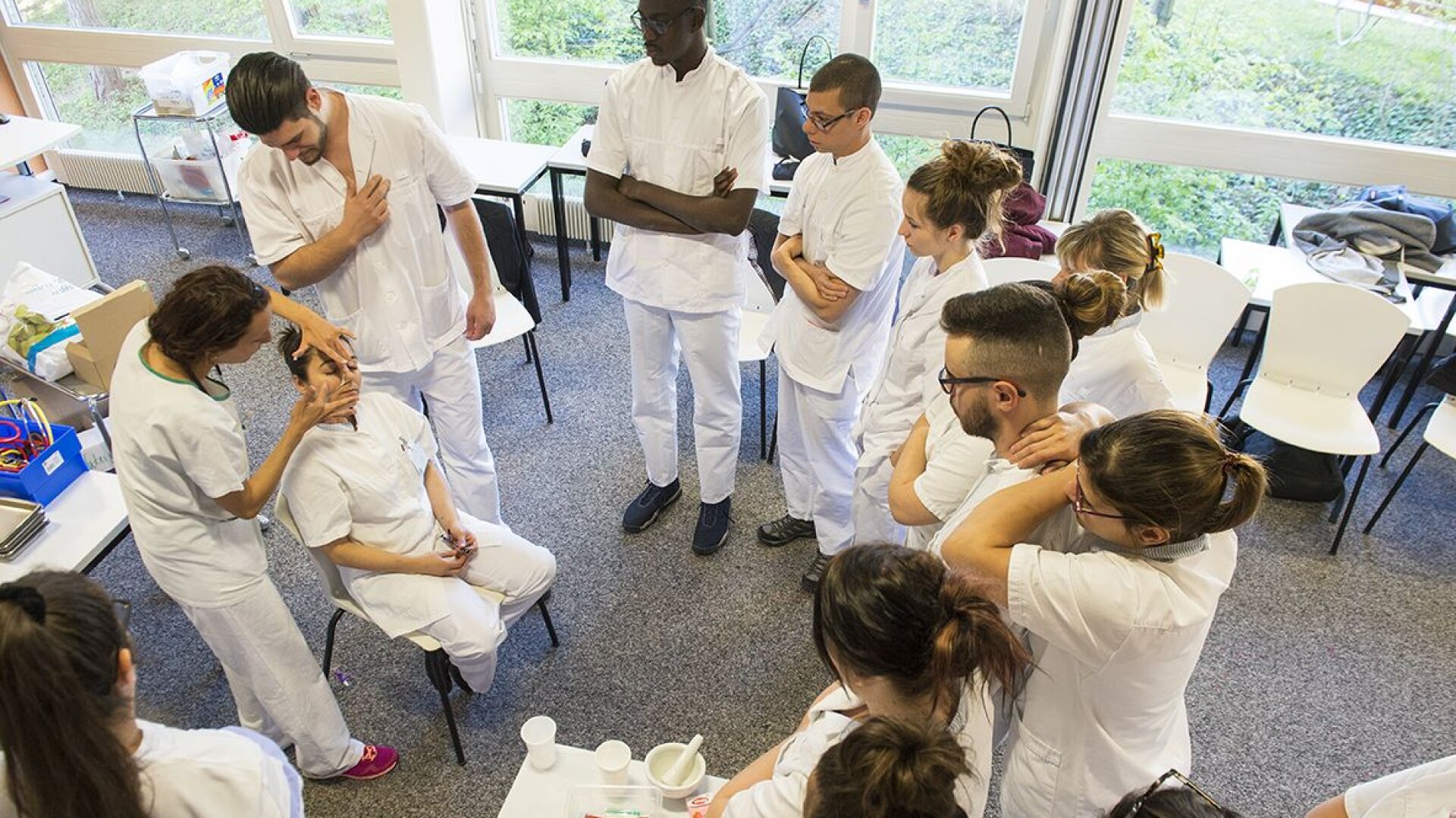 Un groupe d'étudiants en médecine en blouse blanche se réunit autour d'une femme assise pendant qu'un instructeur examine le visage de la femme dans une salle de classe lumineuse remplie de chaises et de tables.