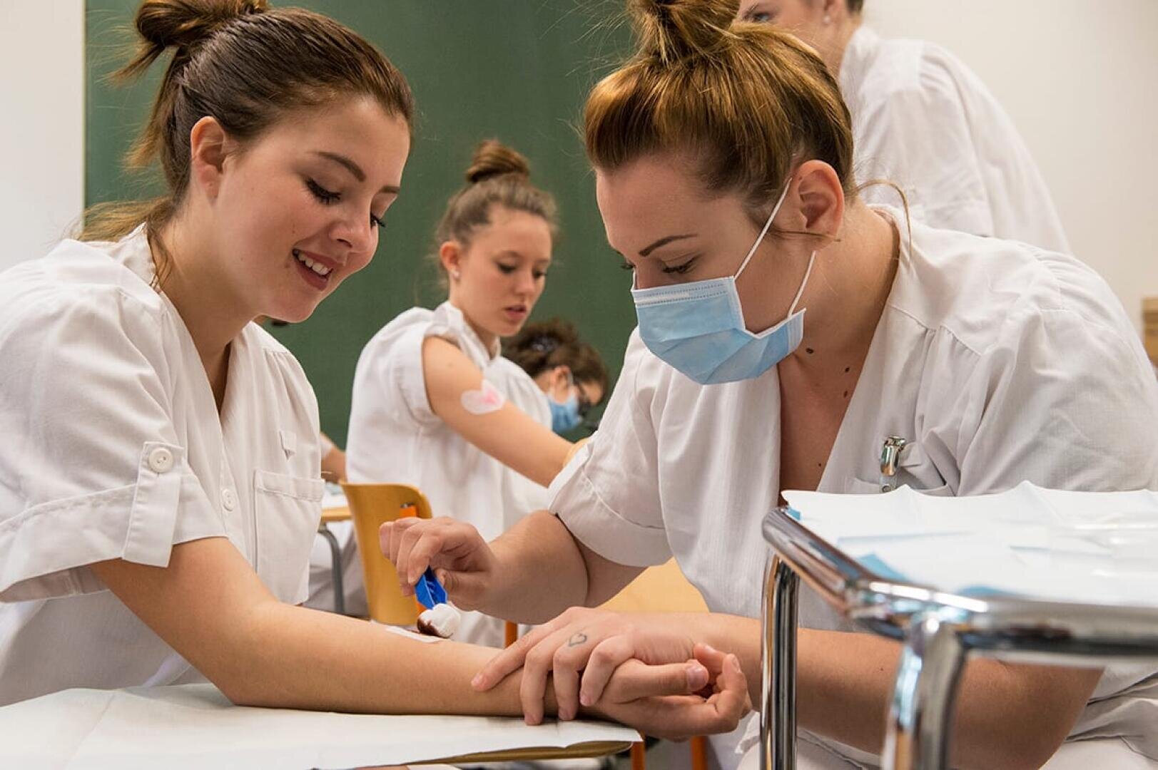 Un agent de santé portant un masque prélève du sang sur le bras d'une jeune femme souriante. Tous deux sont vêtus d'uniformes blancs. À l'arrière-plan, d'autres personnes portant des vêtements similaires sont assises et interagissent.