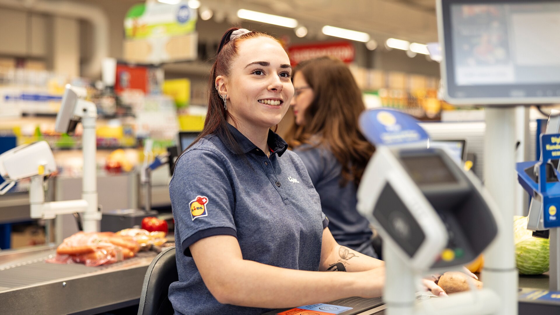 Une caissière souriante en uniforme bleu est assise à la caisse d'un supermarché, avec des produits d'épicerie comme du pain et des légumes sur le tapis roulant et un autre employé à l'arrière-plan.