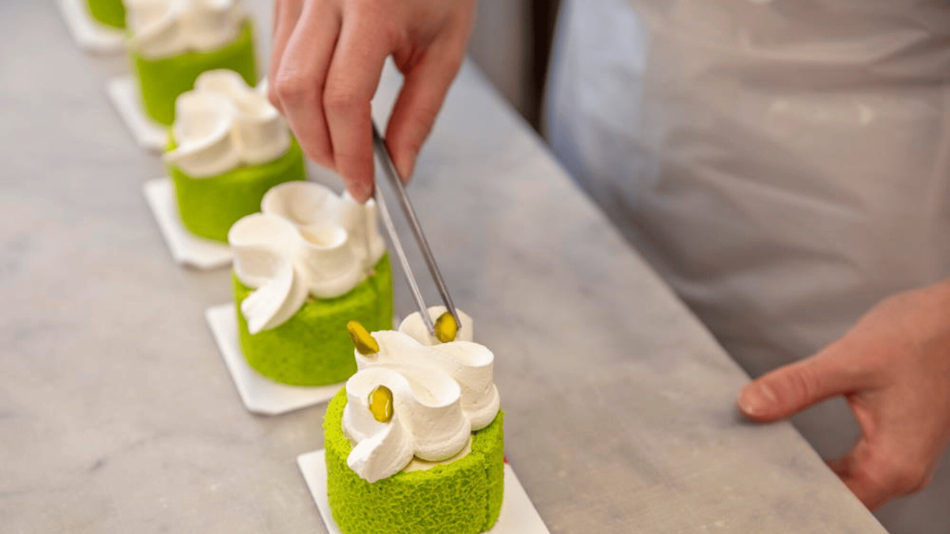 Une personne utilise des pinces pour placer des pistaches sur de petits gâteaux ronds verts garnis de tourbillons de crème fouettée blanche, disposés en rang sur une surface en marbre.