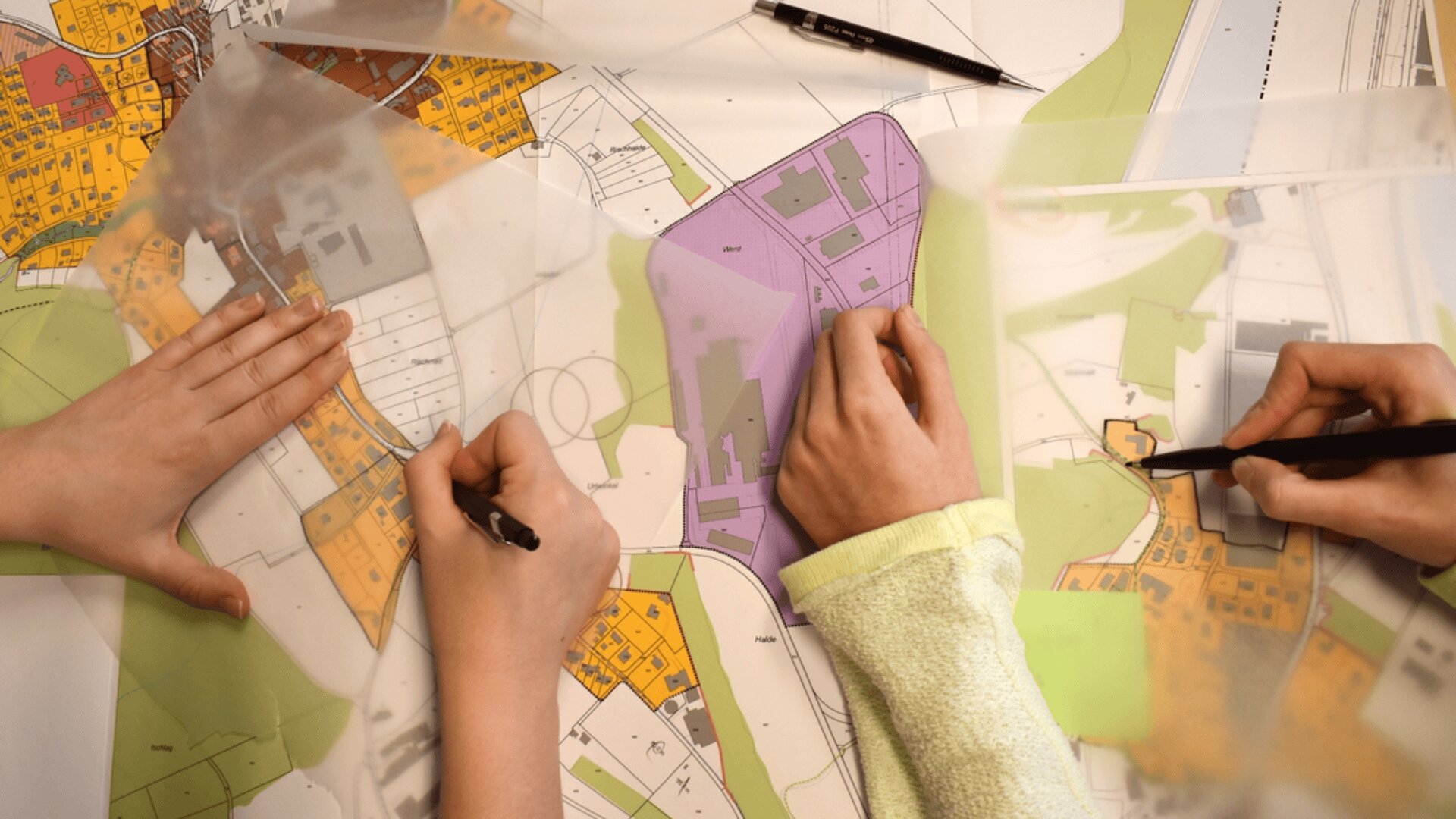 Deux personnes dessinent et marquent sur une carte d'urbanisme colorée, utilisant des stylos et des feuilles transparentes pour superposer des dessins et des notes sur le plan.
