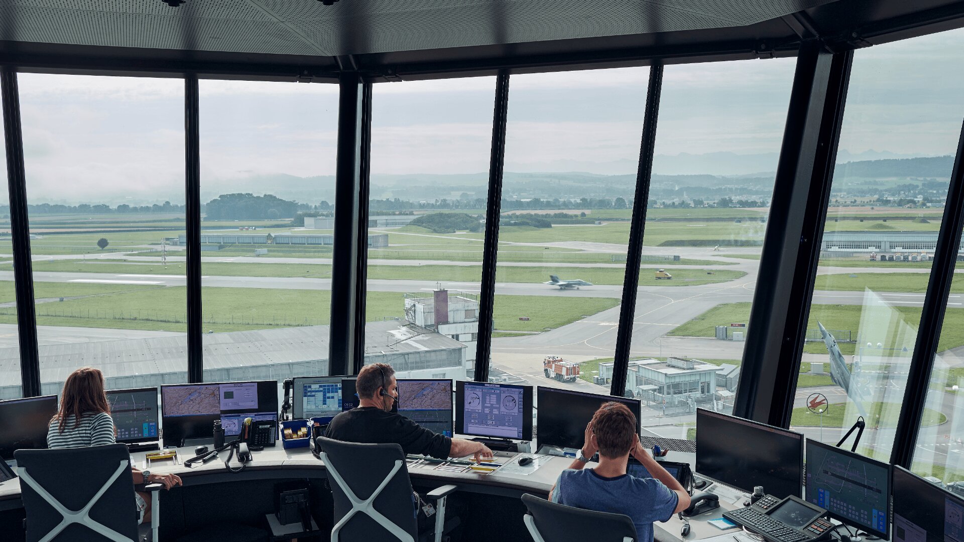 Trois personnes travaillent à des postes informatiques à l'intérieur d'une tour de contrôle d'aéroport, avec vue sur les pistes, les voies de circulation et un avion à travers de grandes fenêtres. L'espace de travail est rempli d'écrans et d'équipements multiples.