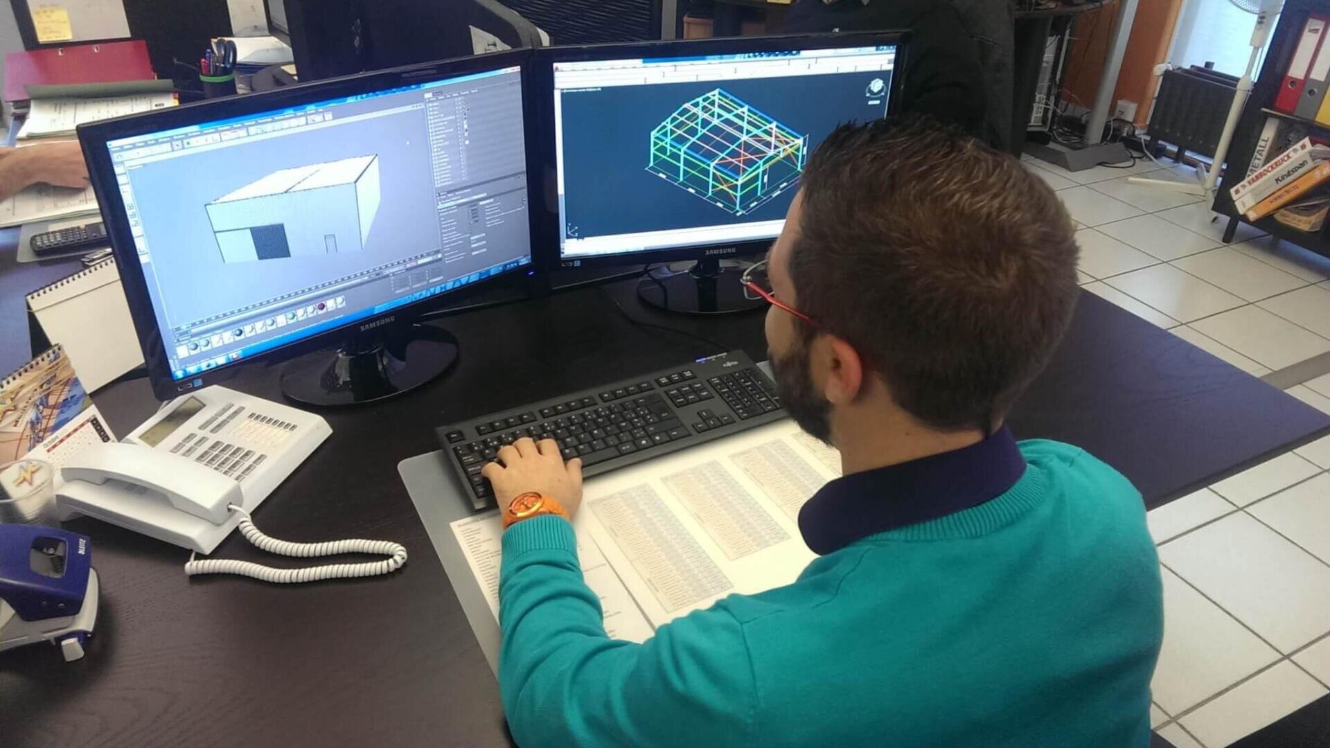 Un homme est assis à un bureau et travaille sur deux écrans d'ordinateur affichant des modèles architecturaux et structurels en 3D. Il utilise une souris et un clavier. Des documents et des fournitures de bureau sont posés sur le bureau autour de lui.