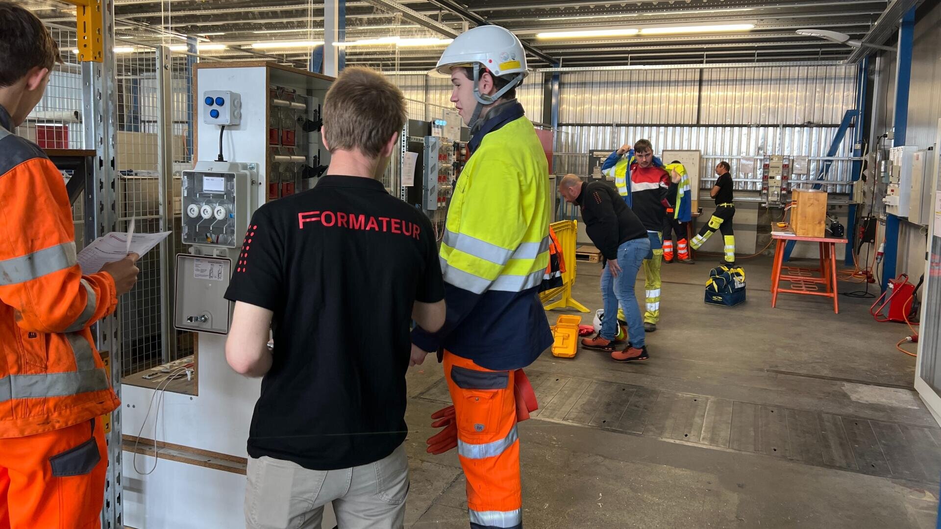 Plusieurs travailleurs en tenue de sécurité se trouvent à l'intérieur d'un grand atelier industriel. L'un d'entre eux porte une chemise FORMATEUR et supervise d'autres personnes portant des gilets et des casques fluorescents, dont certaines interagissent avec des équipements posés sur des tables.