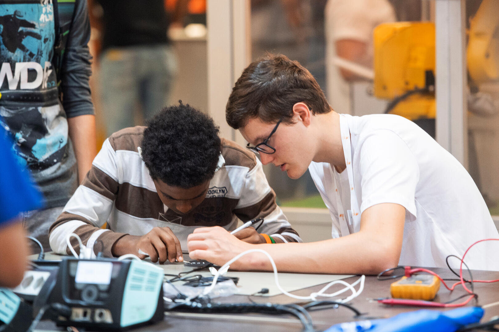 Deux jeunes hommes sont assis à une table et se concentrent sur l'assemblage ou la réparation d'un appareil électronique, entourés d'outils et de fils, dans un atelier ou une salle de classe.