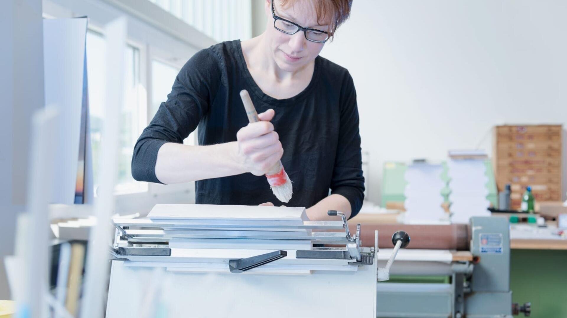 Une personne portant des lunettes et une chemise noire utilise un pinceau pour appliquer de la colle ou de la peinture sur une pile de papier dans un espace de travail moderne et lumineux, avec divers équipements en arrière-plan.