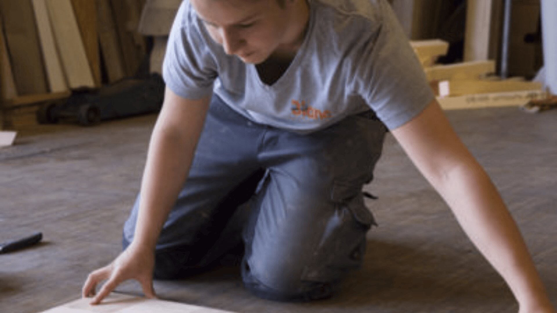 Une personne est agenouillée sur le sol, mesurant ou alignant une planche de bois, vêtue d'une chemise grise et d'un pantalon de travail dans un atelier.