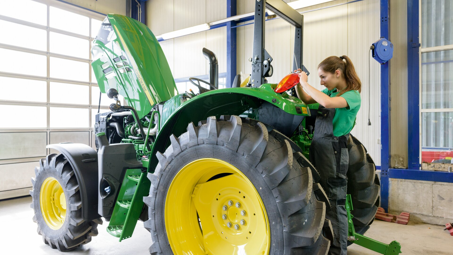 Une femme en bleu de travail inspecte et répare un tracteur vert à l'intérieur d'un atelier bien éclairé, le capot du tracteur étant ouvert et les outils visibles à proximité.