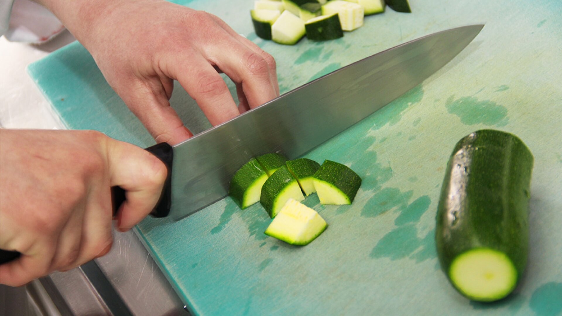 Une personne coupe une courgette en cubes sur une planche à découper verte à l'aide d'un grand couteau de cuisine.