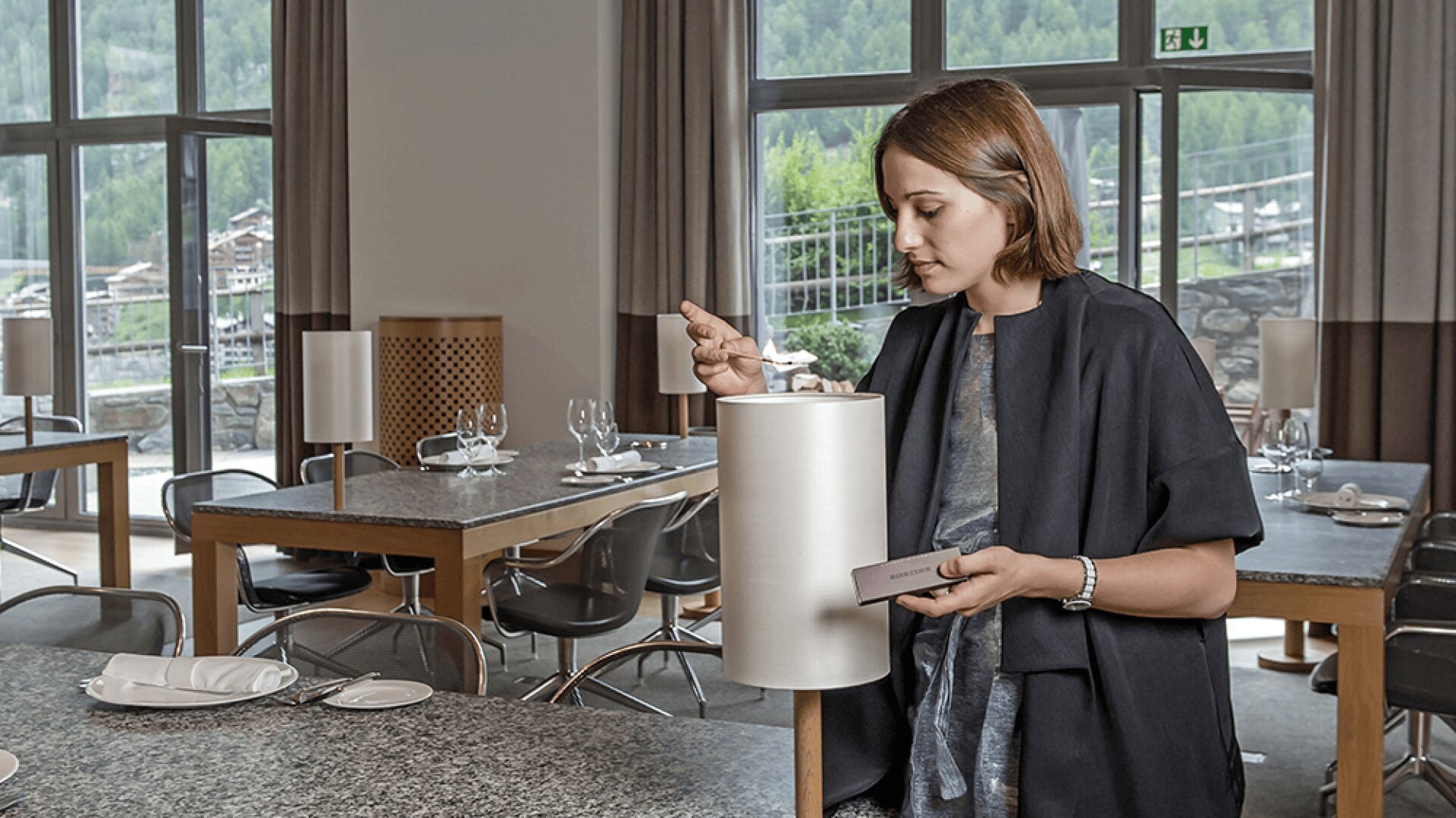Une femme se tient debout dans un restaurant moderne et vide, tenant une petite carte et examinant un plat présenté à l'intérieur d'un grand récipient cylindrique blanc sur une table dressée pour le repas. De grandes fenêtres offrent une vue pittoresque sur l'extérieur.