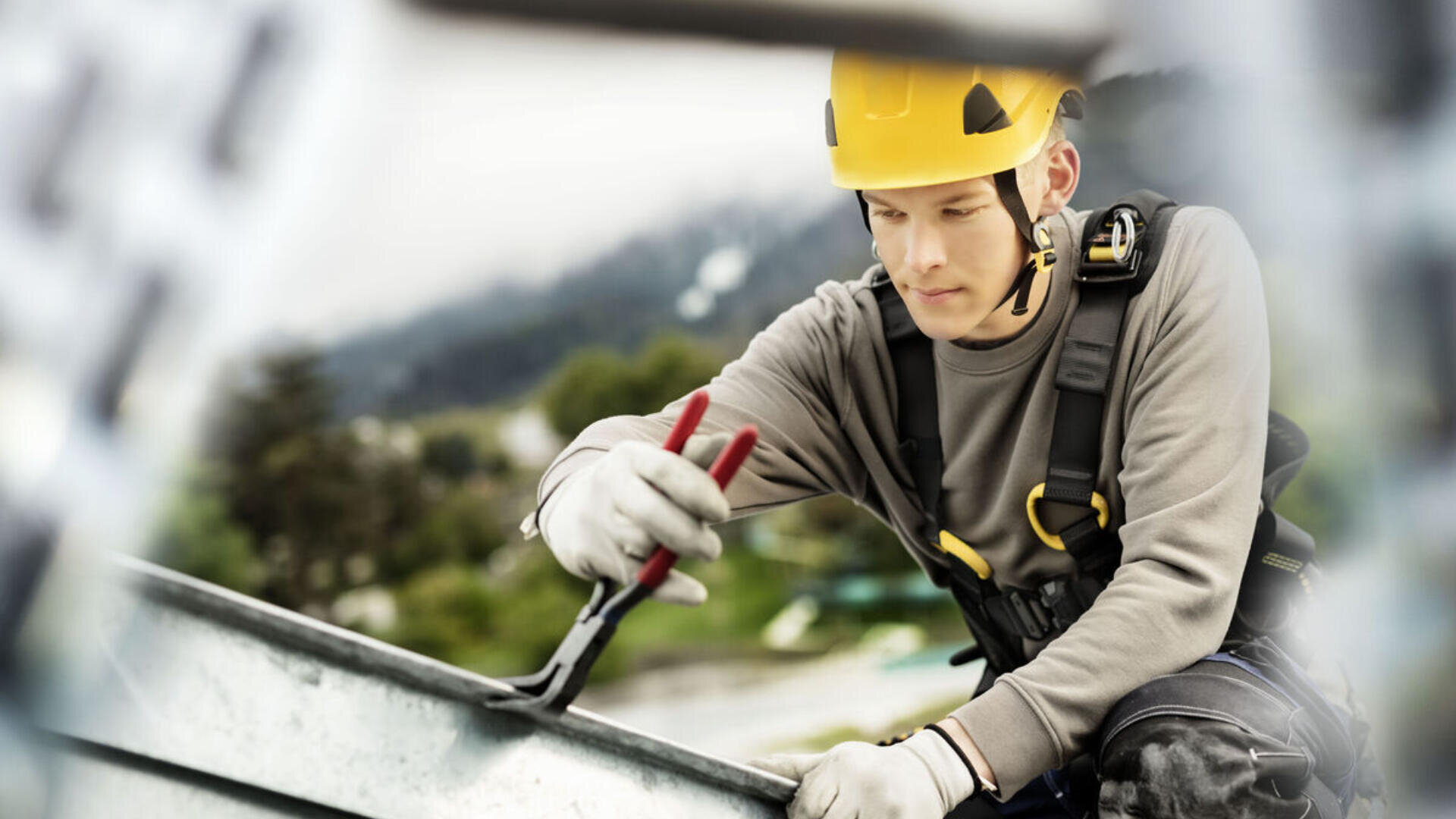 Un ouvrier de la construction portant un casque de sécurité jaune et des gants utilise des pinces tout en travaillant sur une structure métallique en plein air, avec un arrière-plan naturel flou derrière lui.