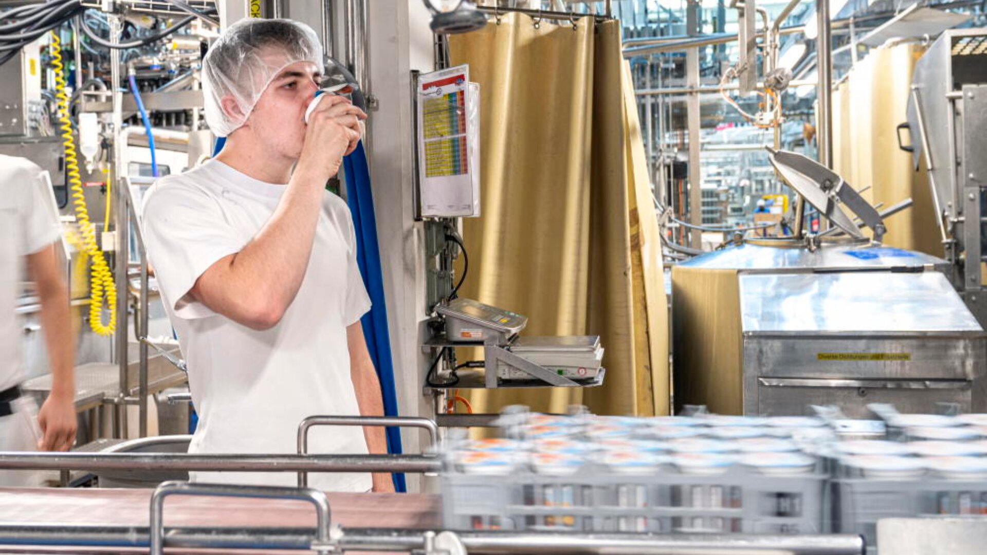 Un ouvrier portant une charlotte et un uniforme blanc boit dans une canette près d'un tapis roulant contenant des produits en conserve dans une usine de production de boissons.
