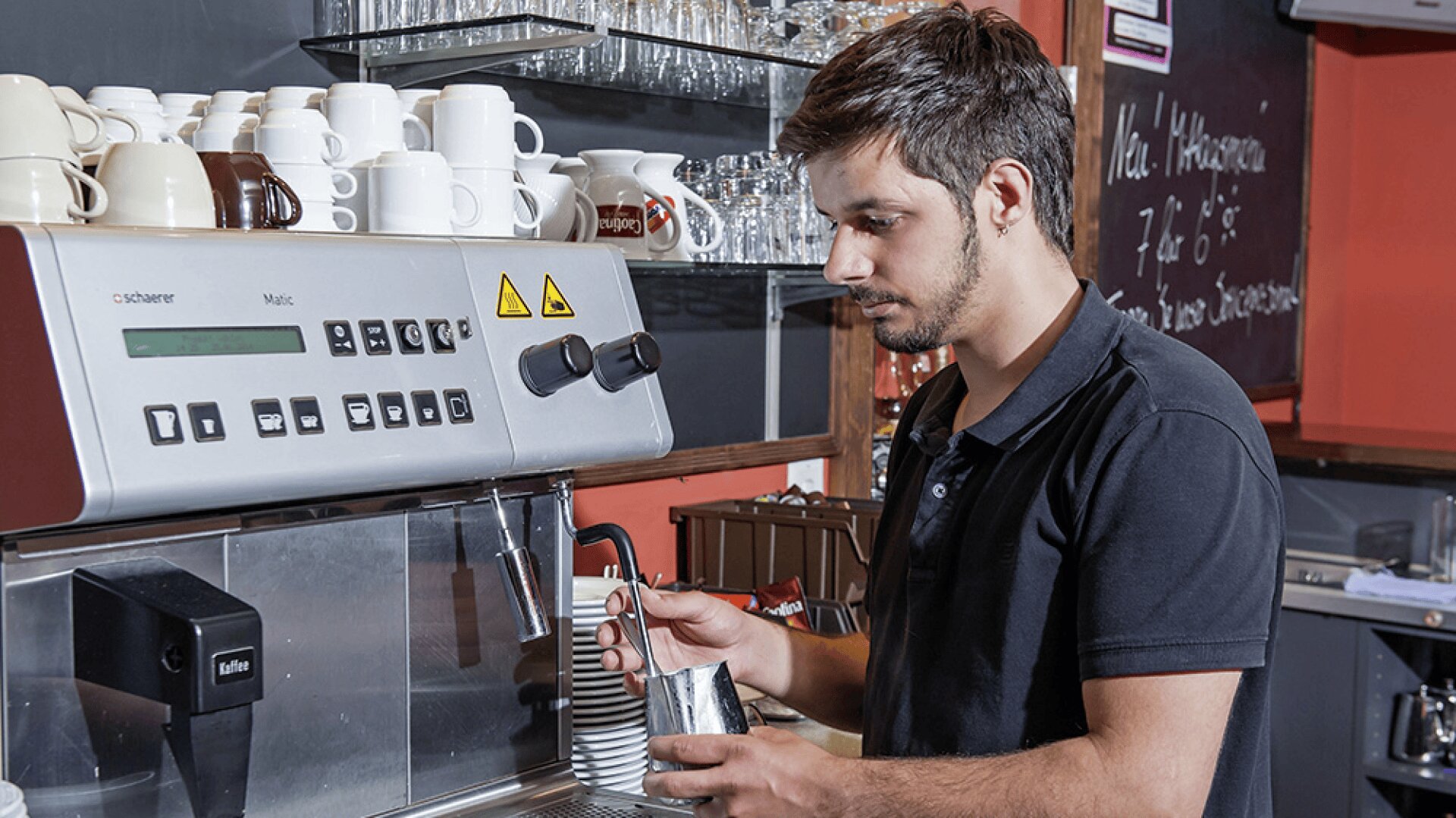 Un barista en chemise noire fait chauffer du lait à l'aide d'une machine à expresso dans un café, avec des étagères de tasses et de verres à l'arrière-plan.
