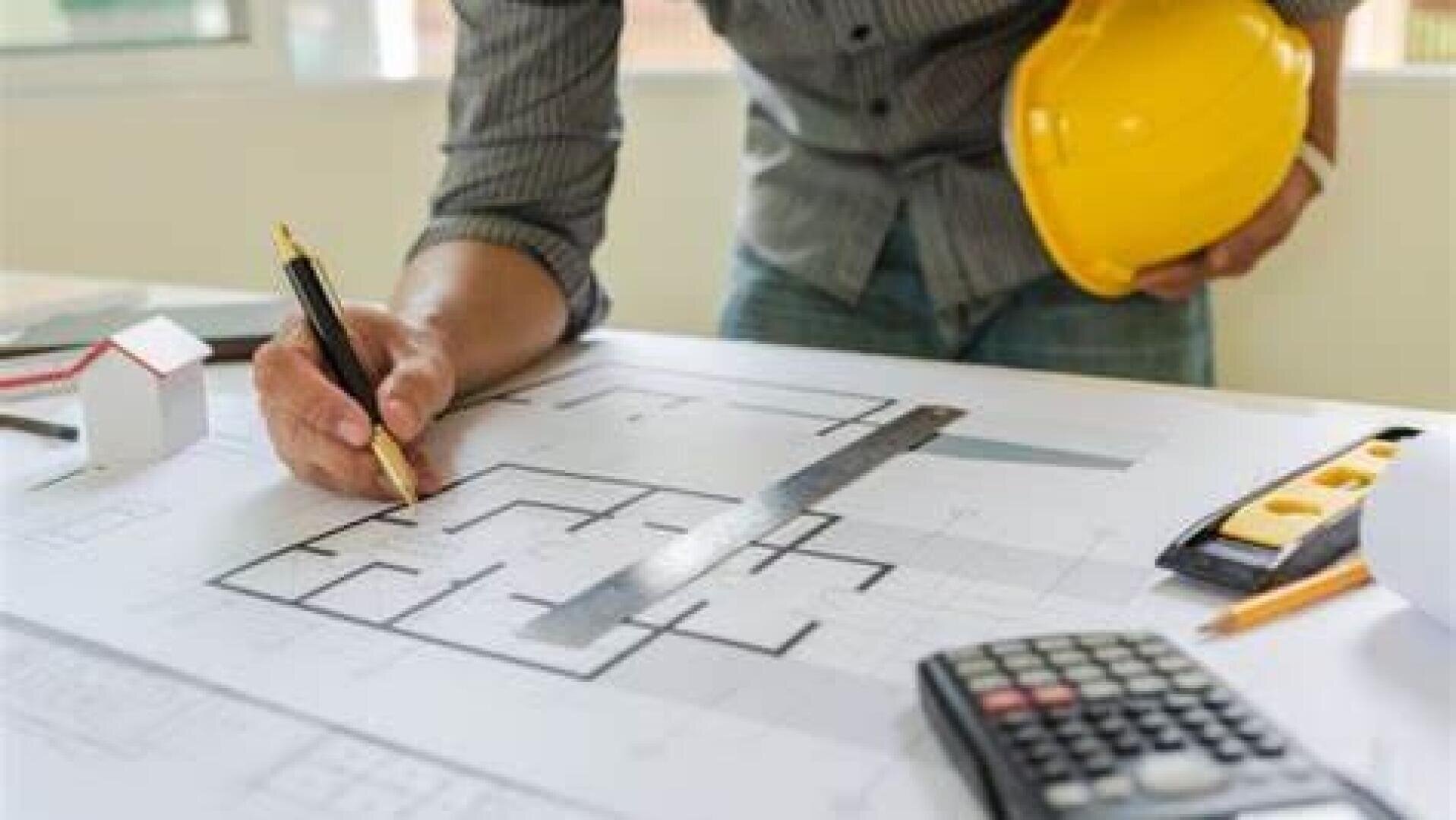 Une personne portant un casque jaune revoit des plans sur un bureau, à l'aide d'une règle et d'un stylo. À proximité se trouvent une calculatrice, une petite maquette de maison et des plans d'architecture, suggérant un travail de construction ou d'architecture.