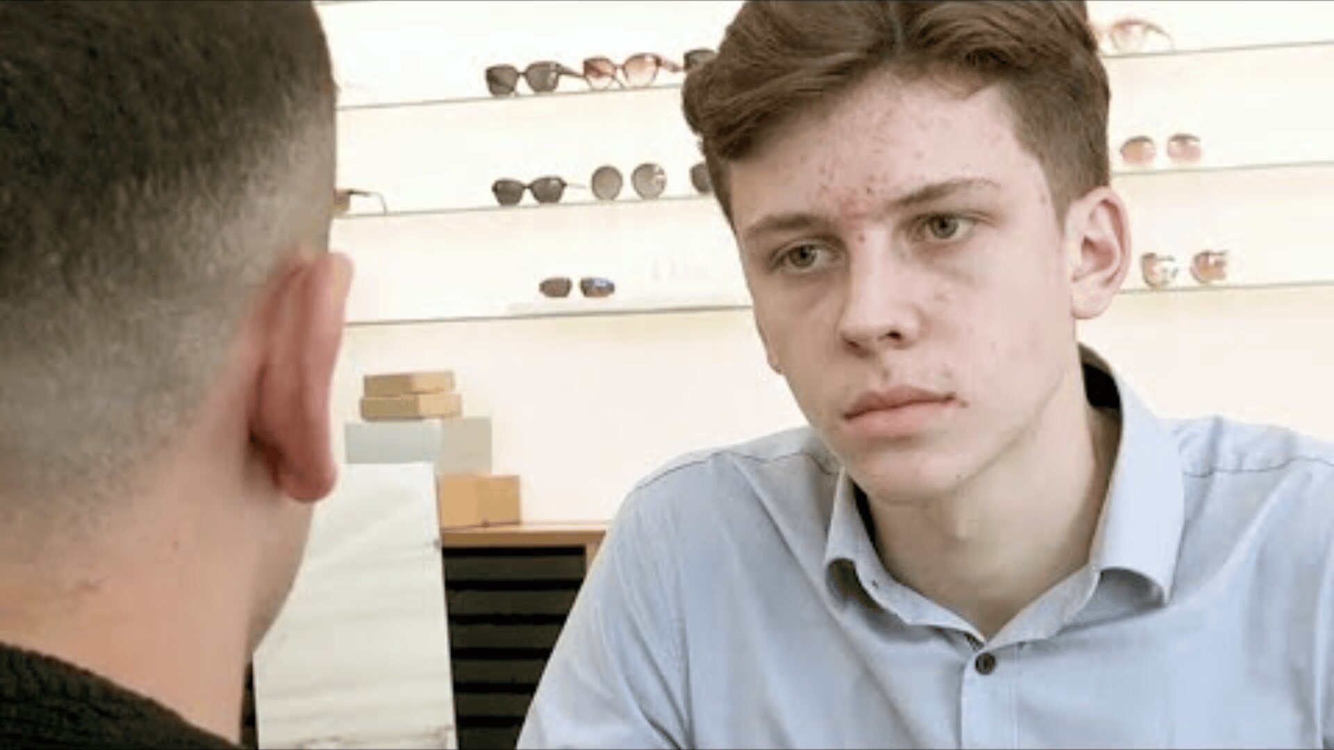 Un jeune homme aux cheveux bruns courts et à l'acné, vêtu d'une chemise bleu clair, est assis à l'intérieur et regarde sérieusement une autre personne qui lui tourne le dos. Des lunettes de soleil sont exposées sur des étagères derrière eux.