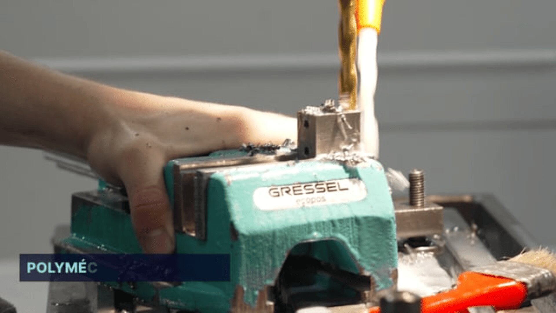 Gros plan d'une main qui maintient une pièce de métal dans un étau Gressel vert, tandis qu'une perceuse s'y enfonce, le liquide de refroidissement ruisselant sur la zone. Le mot POLYMÉC apparaît dans un encadré bleu dans le coin inférieur gauche.