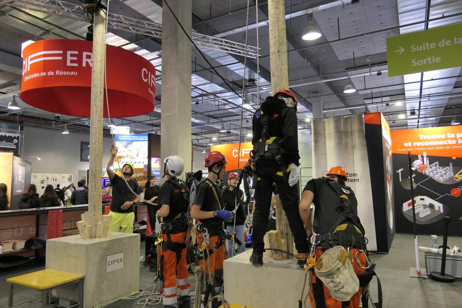 Plusieurs personnes portant des casques et des équipements de sécurité participent à une démonstration d'accrobranche ou d'accès par corde lors d'un événement en intérieur, avec des stands d'information et des panneaux visibles à l'arrière-plan.