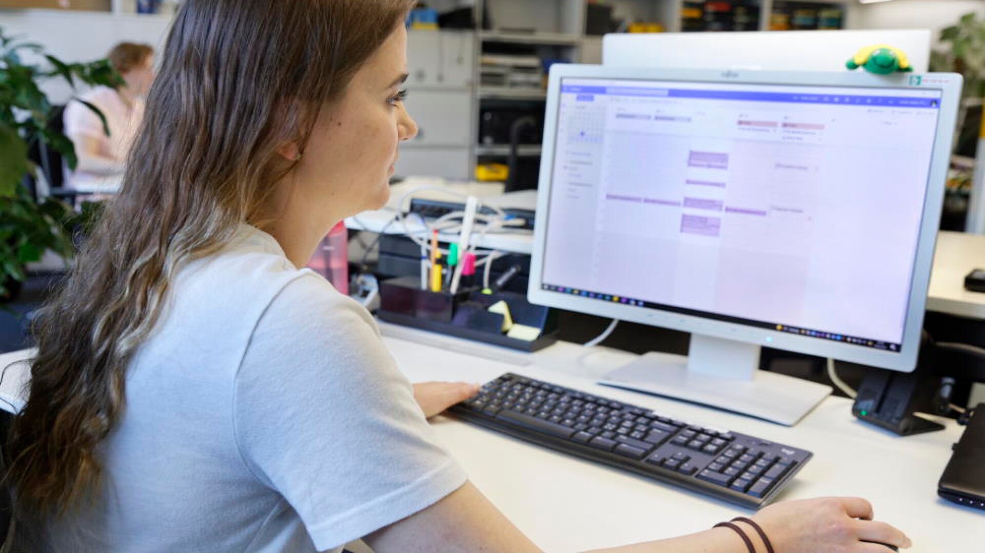 Une femme aux longs cheveux bruns est assise à un bureau, utilisant un ordinateur avec un grand écran affichant une application de planification ou de calendrier. Des fournitures de bureau et des collègues sont visibles à l'arrière-plan.