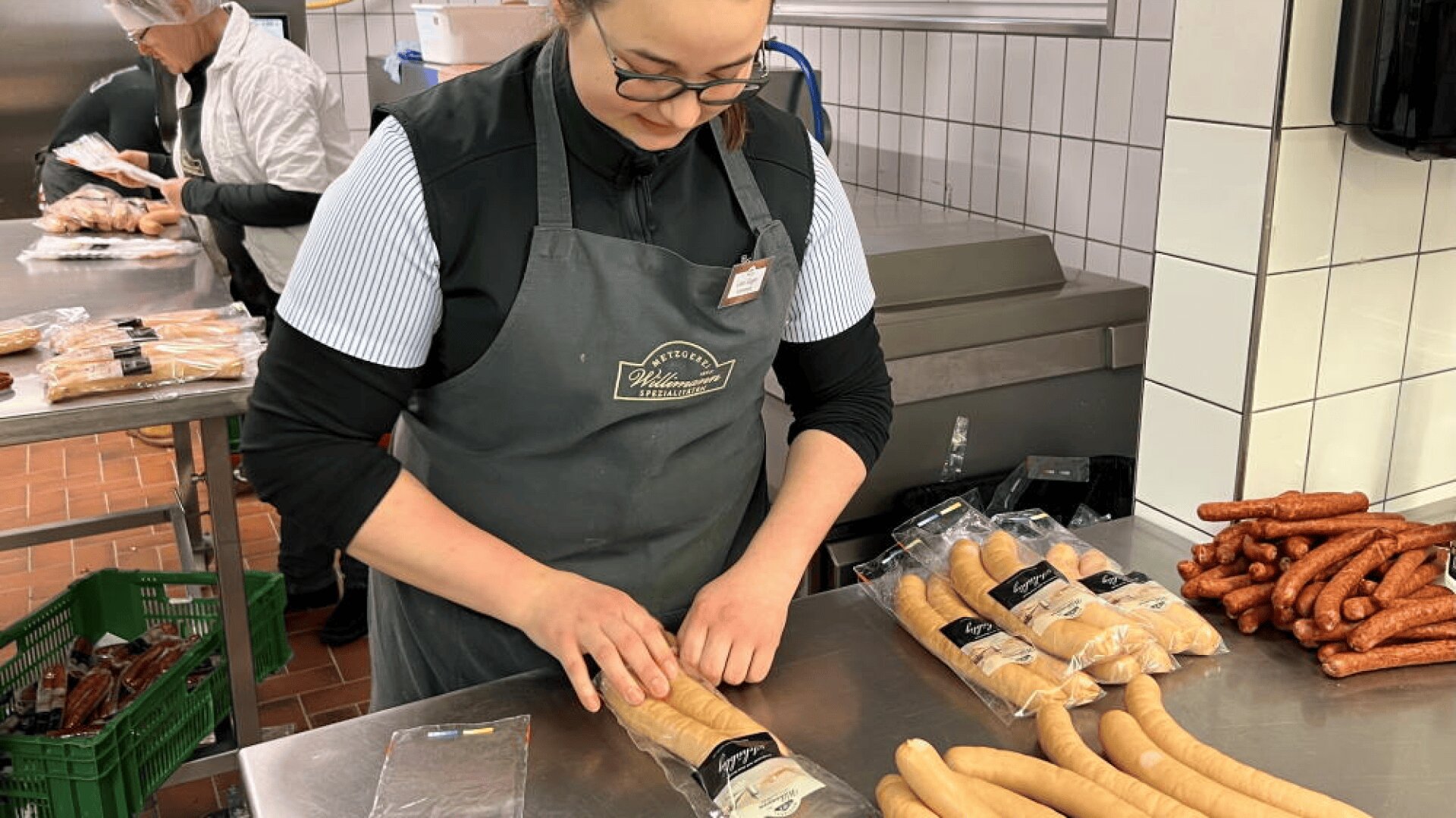 Une femme portant des lunettes et un tablier emballe des saucisses sur un comptoir en acier inoxydable dans une boucherie ou une charcuterie, avec d'autres saucisses et des matériaux d'emballage soigneusement disposés autour d'elle. On aperçoit un autre travailleur à l'arrière-plan.