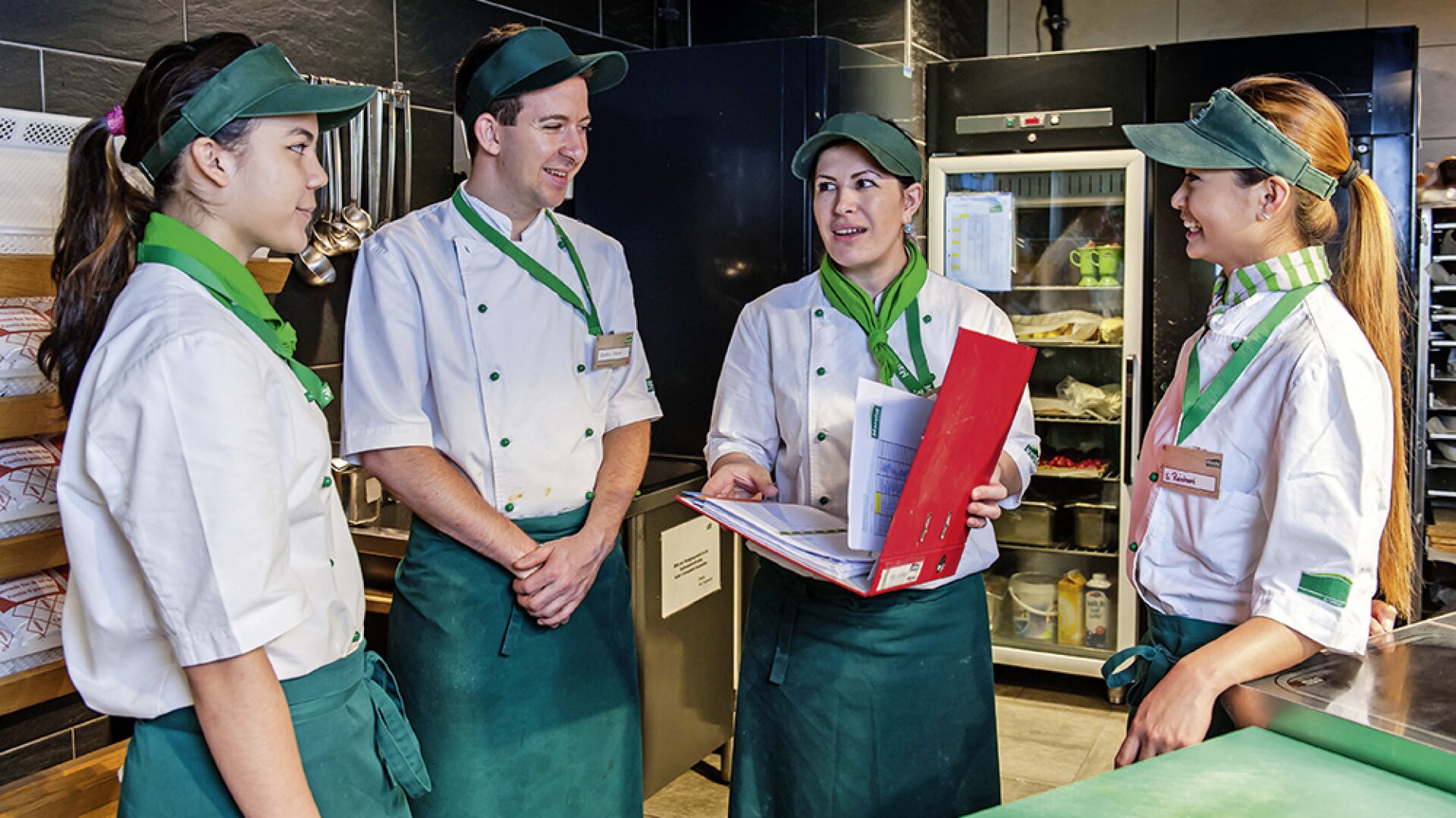 Quatre employés de cuisine en uniforme se tiennent ensemble dans une cuisine commerciale, souriant et discutant. Une personne tient un classeur rouge ouvert, tandis que des étagères et des équipements sont visibles à l'arrière-plan.