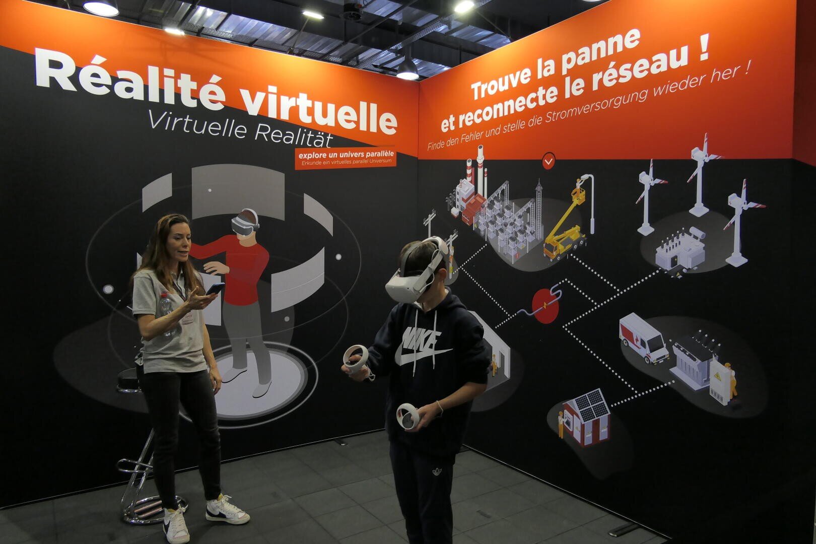Un jeune garçon portant un casque de réalité virtuelle et tenant des manettes interagit avec une exposition de réalité virtuelle sur les réseaux électriques, tandis qu'une femme se tient à proximité dans un stand avec des textes en français et en allemand sur l'énergie et la technologie.