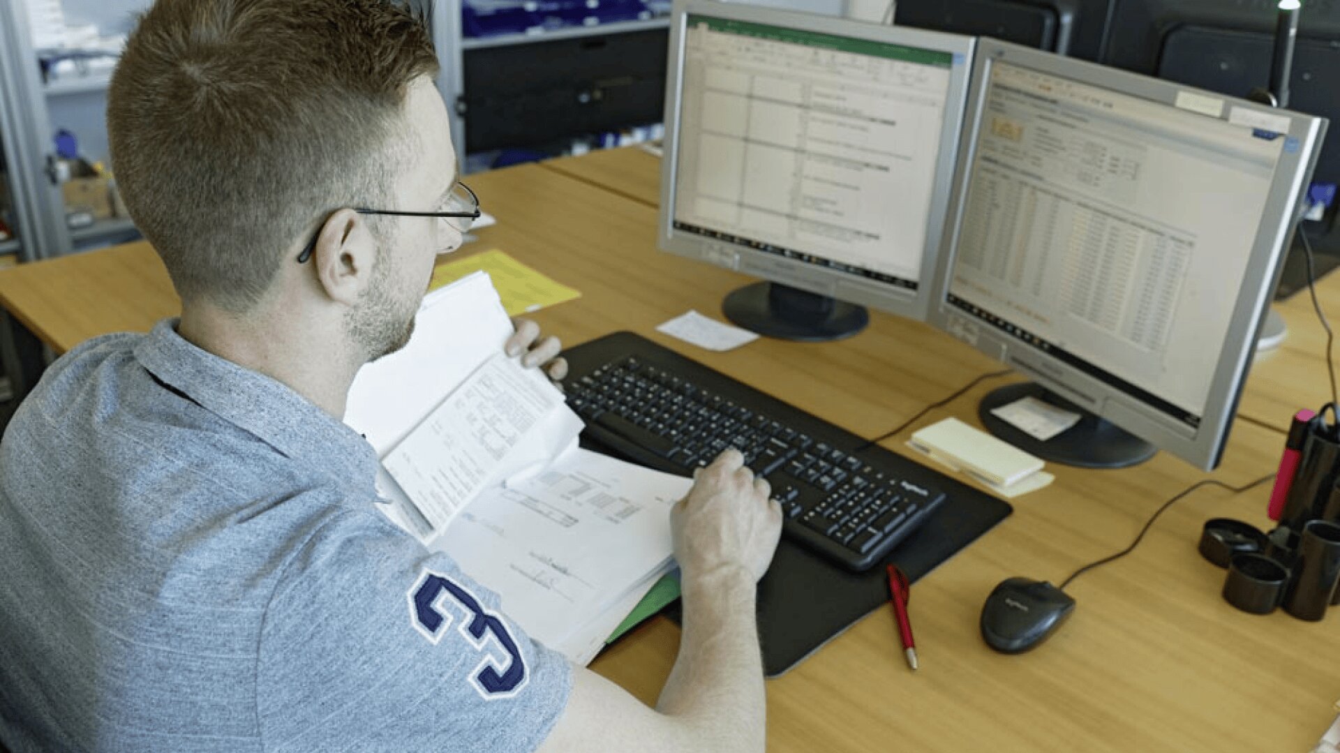 Un homme portant des lunettes et une chemise grise travaille à un bureau où deux écrans d'ordinateur affichent des feuilles de calcul. Il tient un dossier ouvert contenant des papiers et semble examiner des documents.