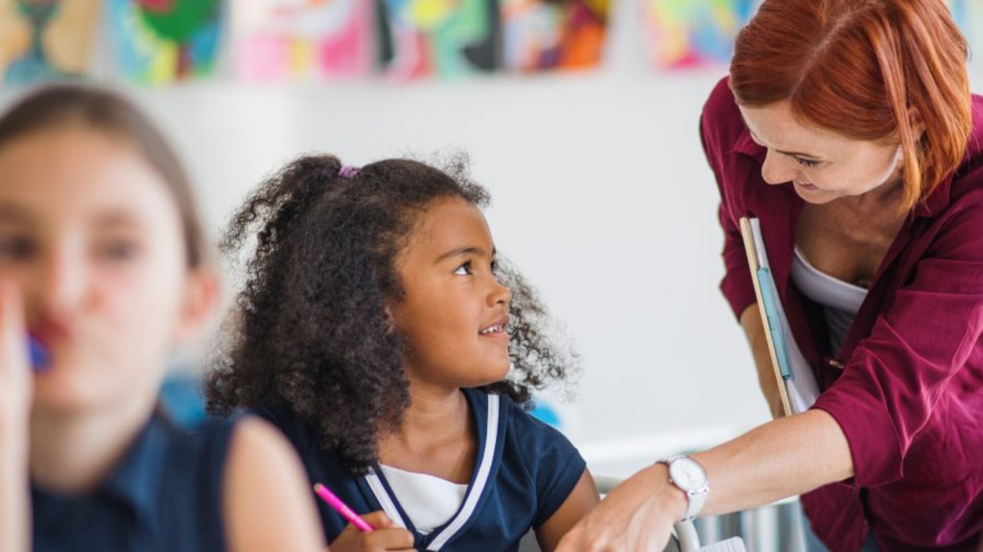 Une jeune fille aux cheveux bouclés sourit et regarde une femme aux cheveux roux qui se penche sur elle et lui parle dans une salle de classe. Un autre élève est flou au premier plan. Des œuvres d'art colorées sont accrochées au mur à l'arrière-plan.