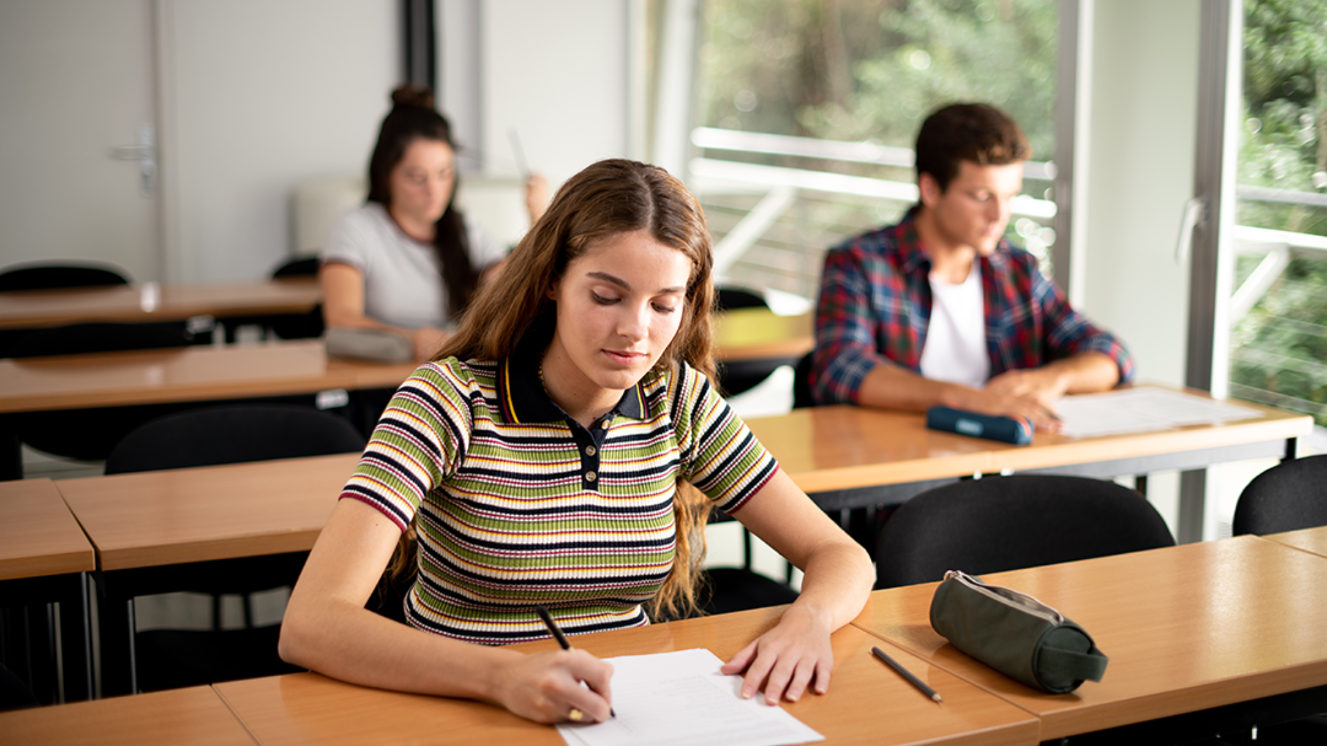 Une jeune femme aux longs cheveux bruns écrit sur un papier à un bureau dans une salle de classe, avec deux autres élèves à l'arrière-plan. La pièce est lumineuse, avec de grandes fenêtres et des bureaux en bois.