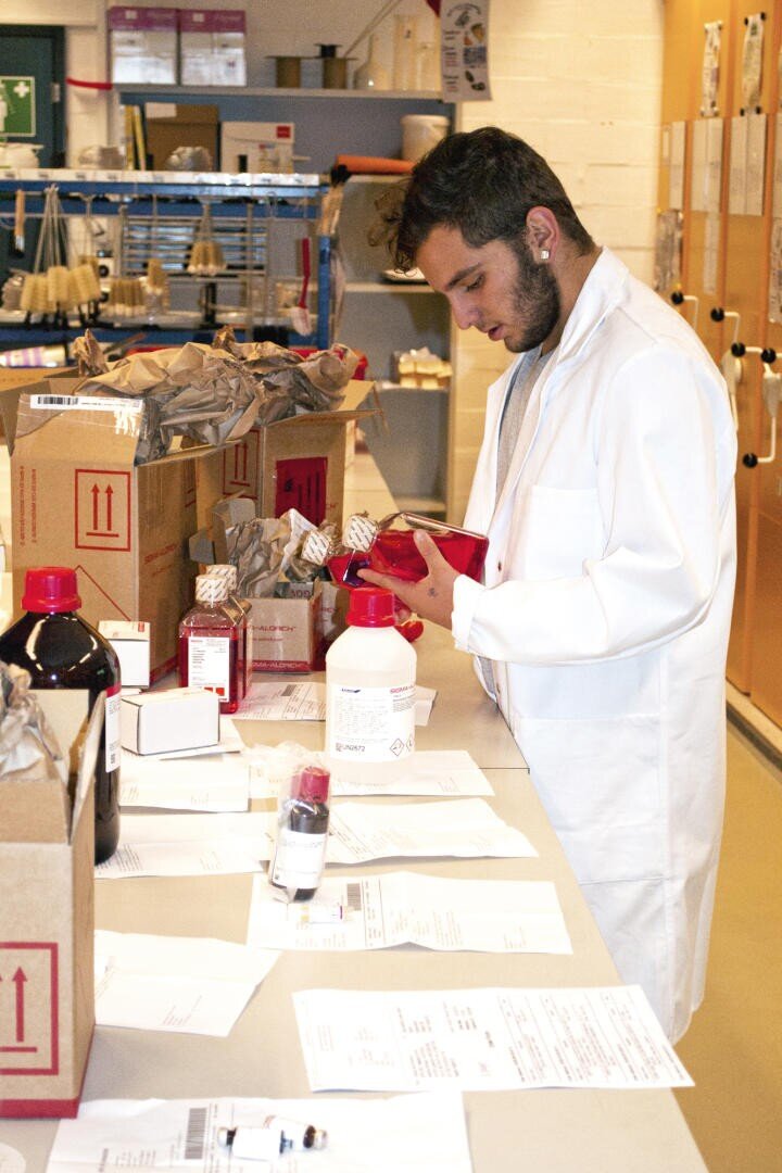 Une personne en blouse blanche examine un flacon de produit chimique rouge sur une paillasse couverte de boîtes, de flacons et de papiers dans un laboratoire.