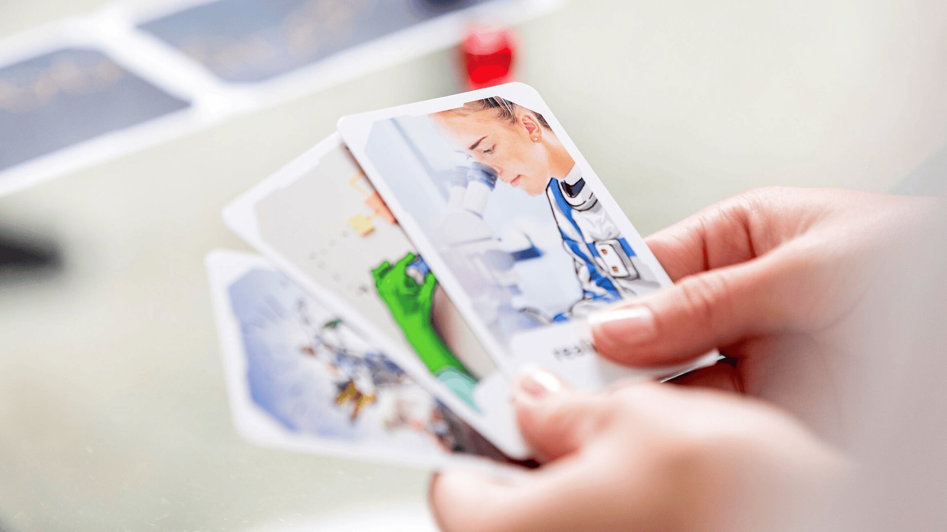 Une personne tient plusieurs cartes dans ses mains. La carte visible montre un scientifique regardant dans un microscope. Les cartes présentent des illustrations colorées et détaillées liées à la science et à la technologie.