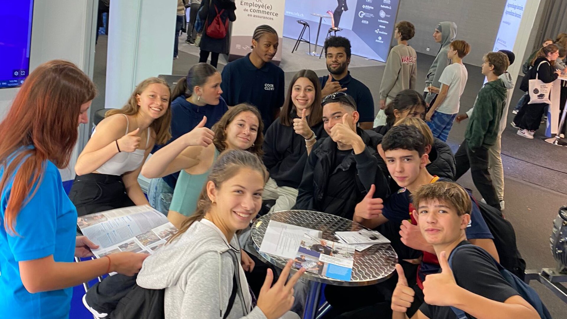 Un groupe d'adolescents souriants est assis et debout autour d'une petite table ronde, levant le pouce devant la caméra. Certains tiennent des brochures et des papiers. Le décor semble être celui d'un événement ou d'une foire en intérieur, avec des banderoles en arrière-plan.