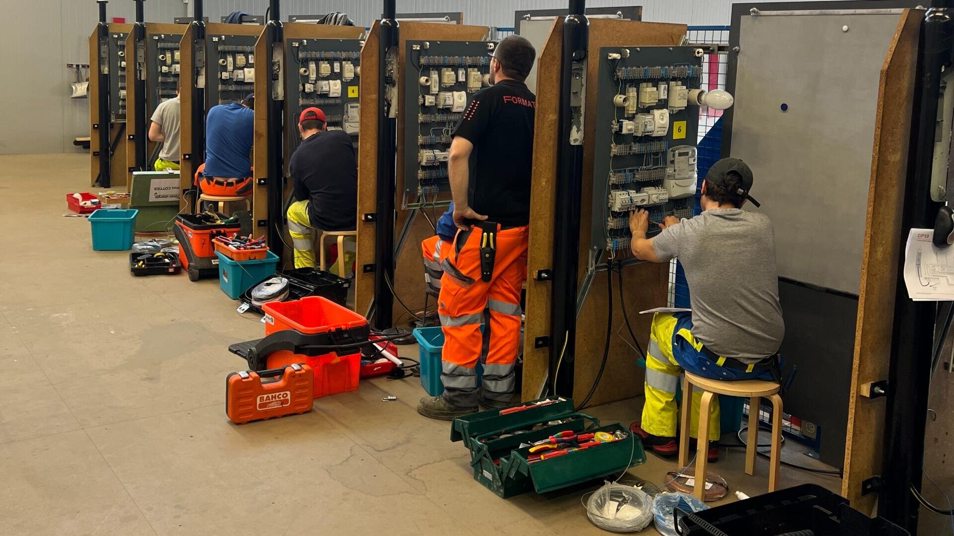 Plusieurs personnes en tenue de travail sont assises ou debout à des postes de travail individuels avec des panneaux électriques, des outils et des boîtes d'équipement dans un atelier.
