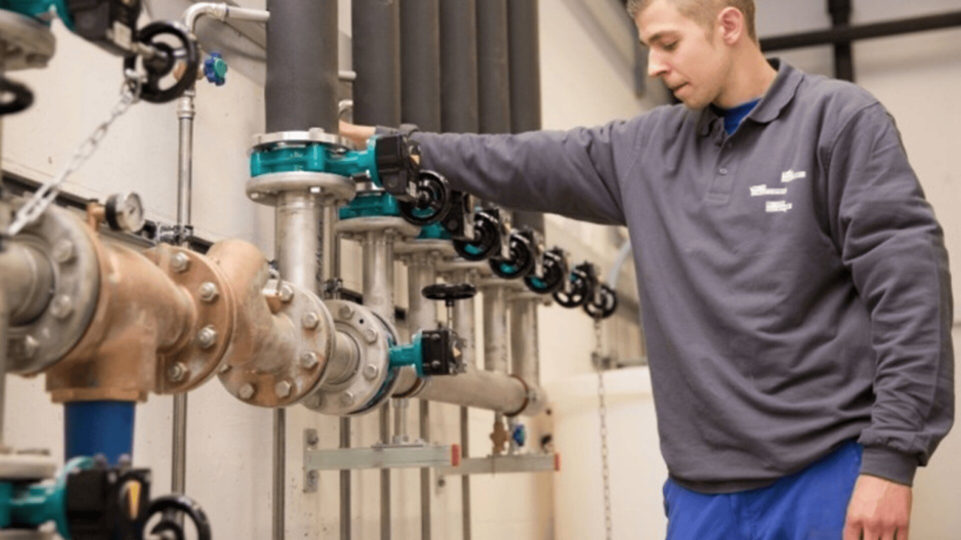 Un homme vêtu d'une chemise grise et d'un pantalon bleu actionne des vannes sur une série de tuyaux industriels montés sur un mur dans un local technique.