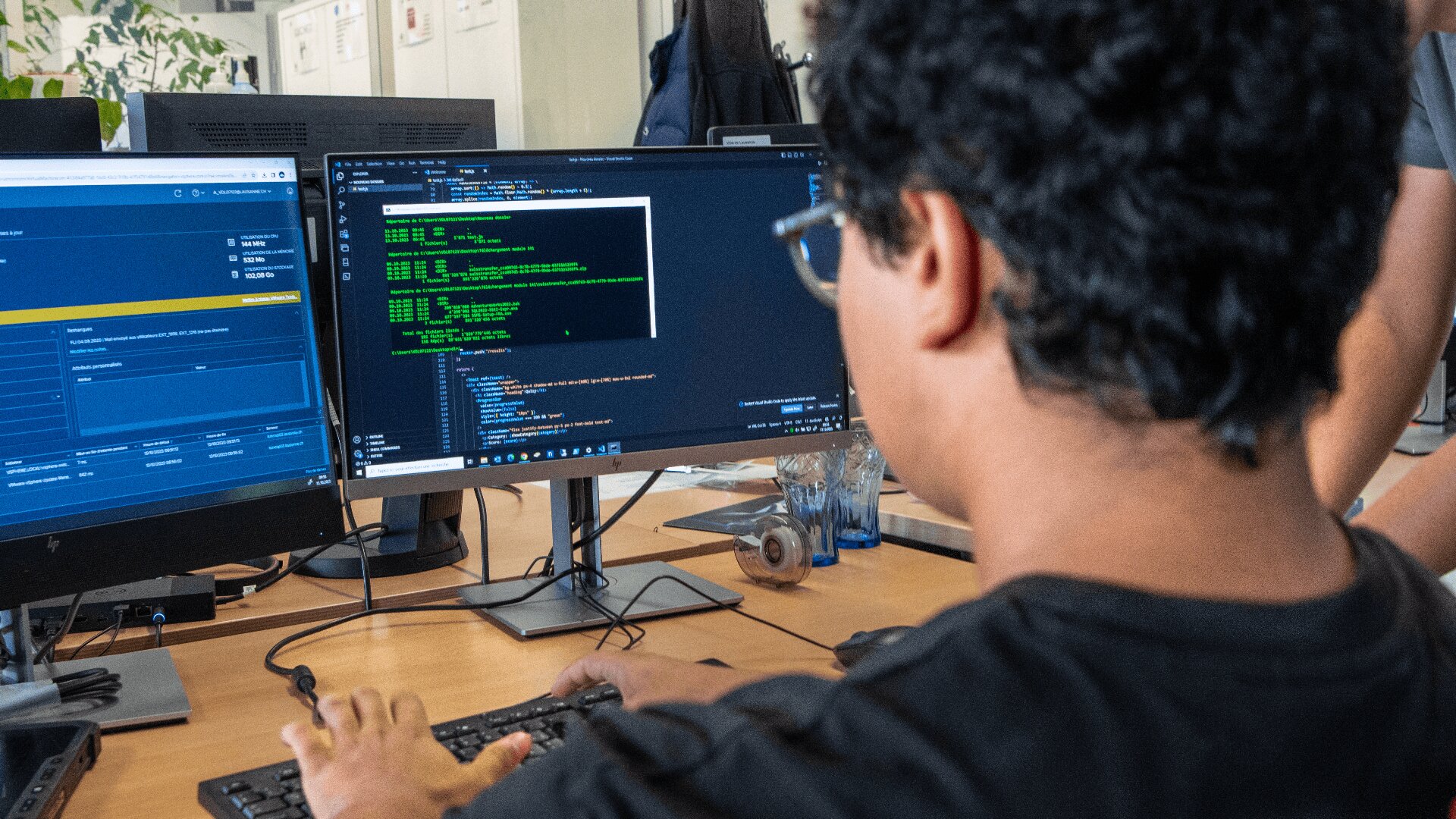 Une personne portant des lunettes est assise à un bureau, tapant sur un clavier et regardant des écrans d'ordinateur affichant des codes et des données dans un bureau moderne.