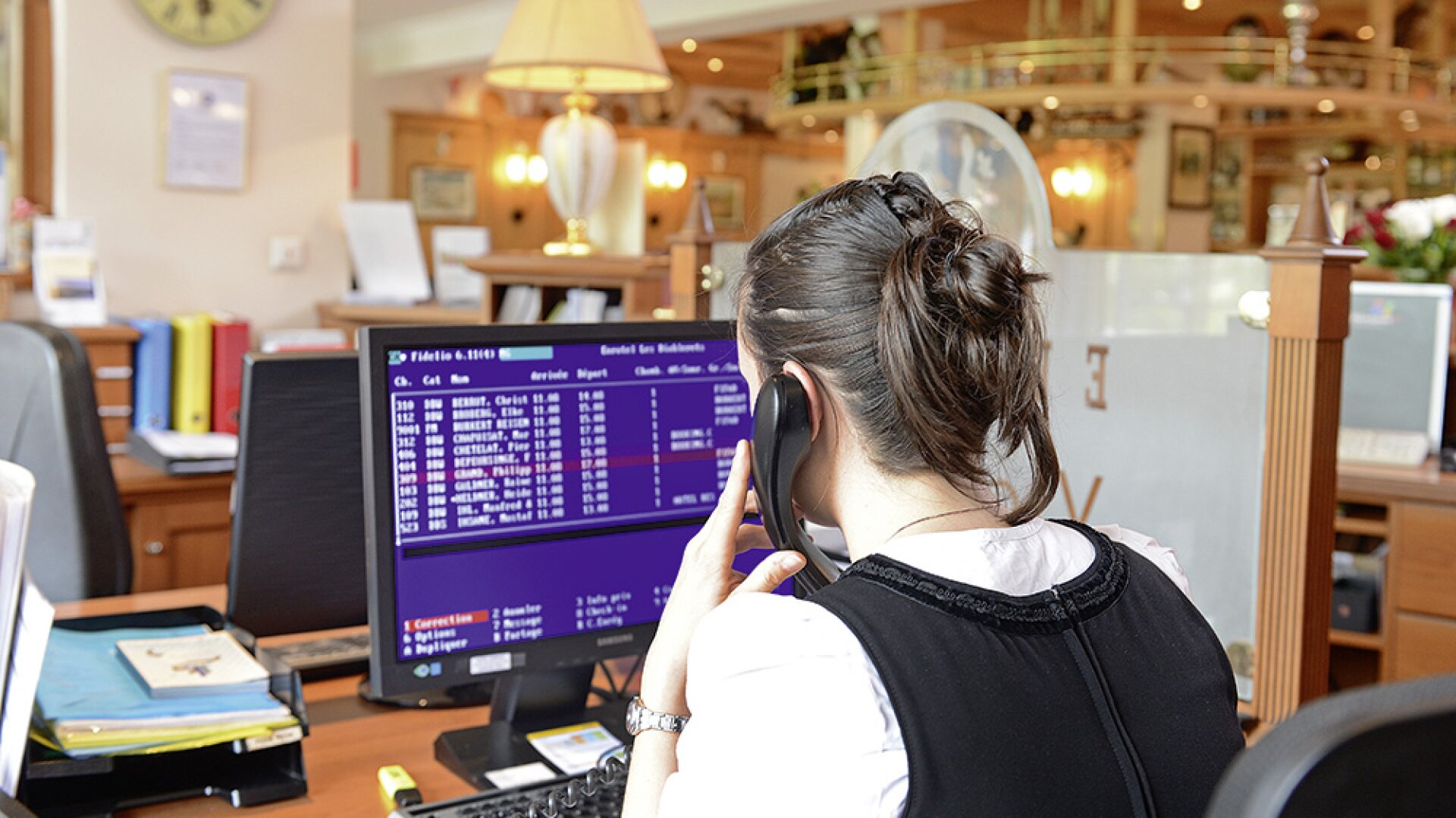 Une femme assise à un bureau dans un bureau ou à la réception d'un hôtel, parlant au téléphone tout en regardant un écran d'ordinateur affichant des informations et des réservations.