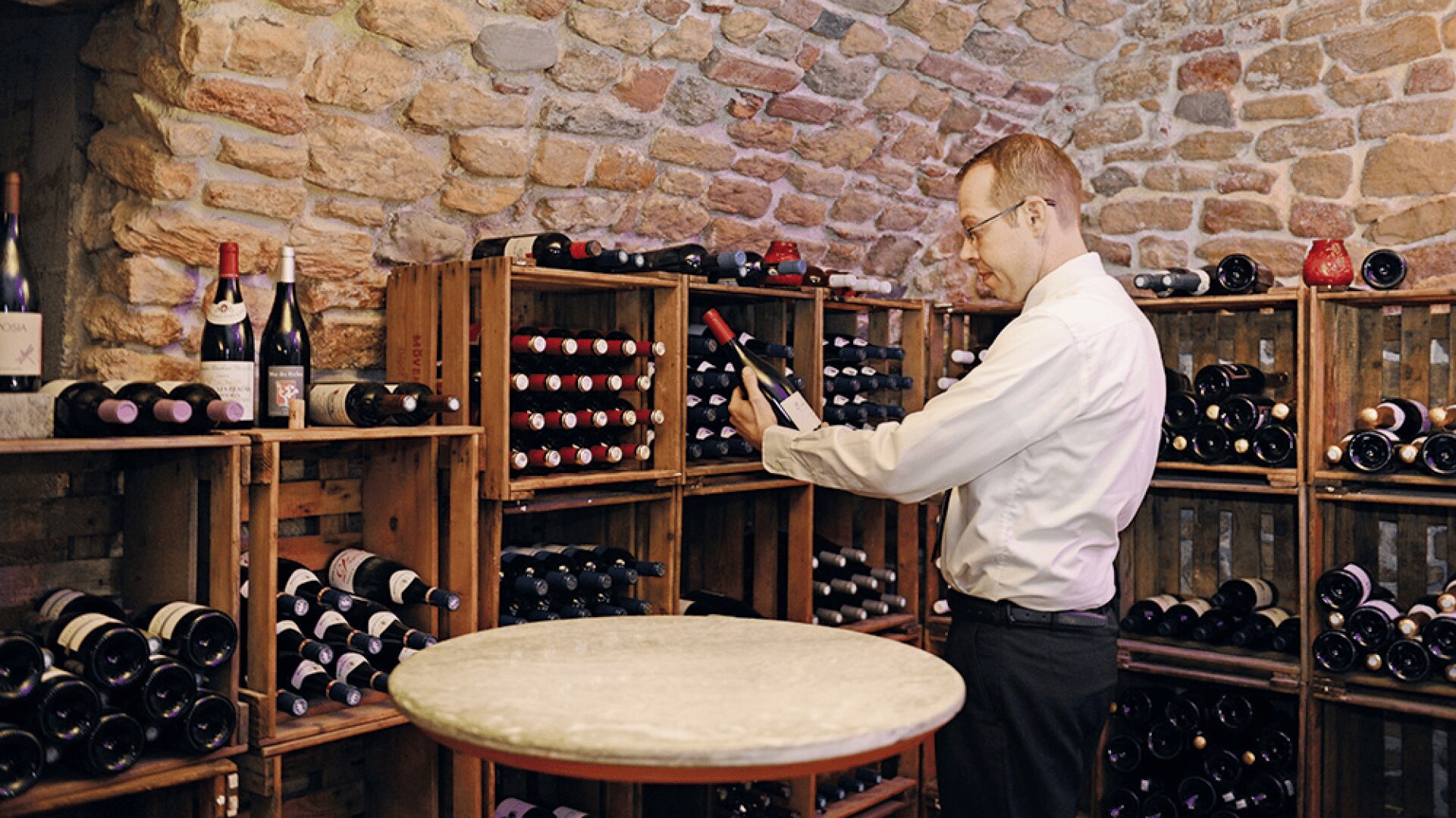 Un homme en chemise blanche examine une bouteille de vin dans une cave à vin rustique avec des murs en pierre et des étagères en bois remplies de diverses bouteilles de vin. Une table ronde en marbre se trouve au premier plan.