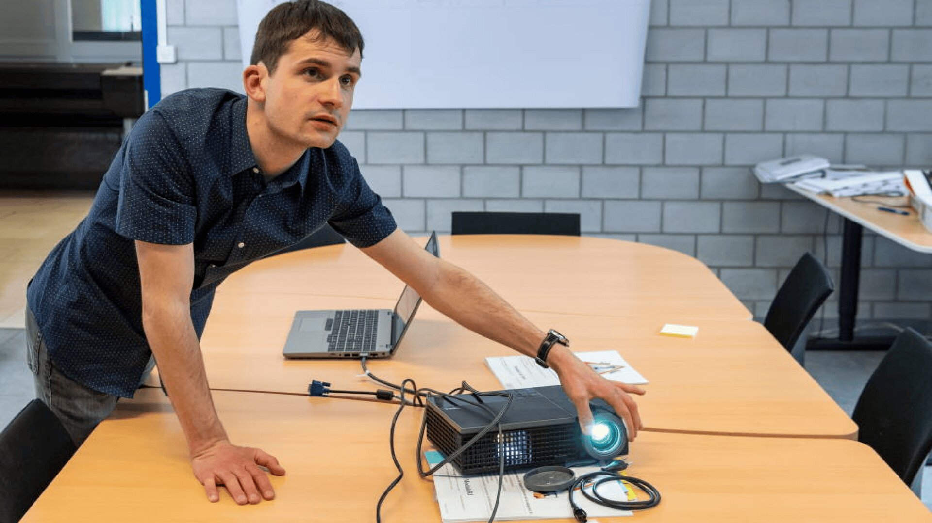 Un homme vêtu d'une chemise à manches courtes se tient près d'une table de conférence, ajustant un projecteur tout en regardant devant lui. Un ordinateur portable et des câbles sont posés sur la table dans un bureau moderne aux murs de briques grises.