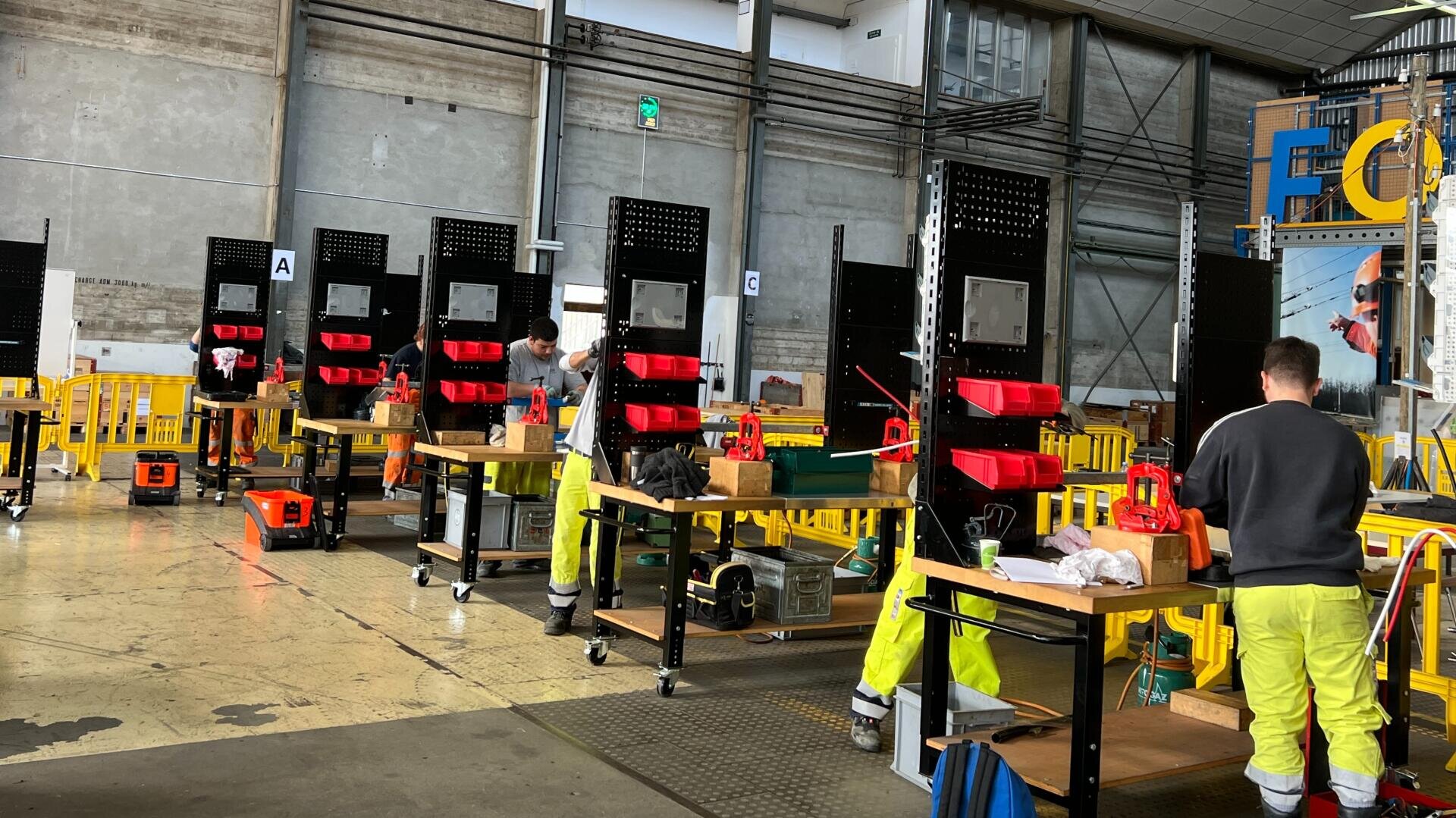 Plusieurs travailleurs en tenue de sécurité assemblent des équipements à des postes de travail distincts dans un atelier industriel doté de murs en béton, de barrières jaunes et d'outils rangés dans des conteneurs rouges.