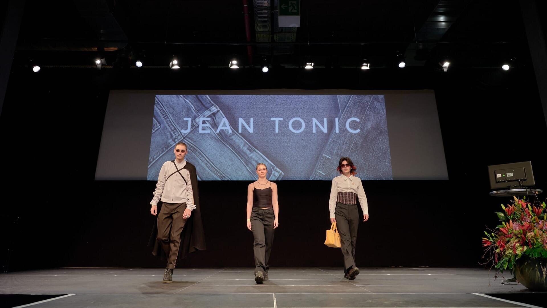 Trois mannequins marchent sur une piste sous des lumières vives, avec un écran derrière eux affichant JEAN TONIC. Elles portent des tenues modernes et élégantes, et des fleurs sont disposées sur le côté de la scène.