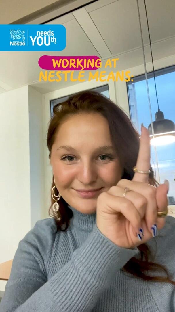 Une femme aux longs cheveux bruns, portant un col roulé gris, sourit et pointe le doigt vers le haut. Le texte indique WORKING AT NESTLÉ MEANS : avec un logo Nestlé et needs YOUth dans le coin. Elle est à l'intérieur, sous une lampe suspendue.
