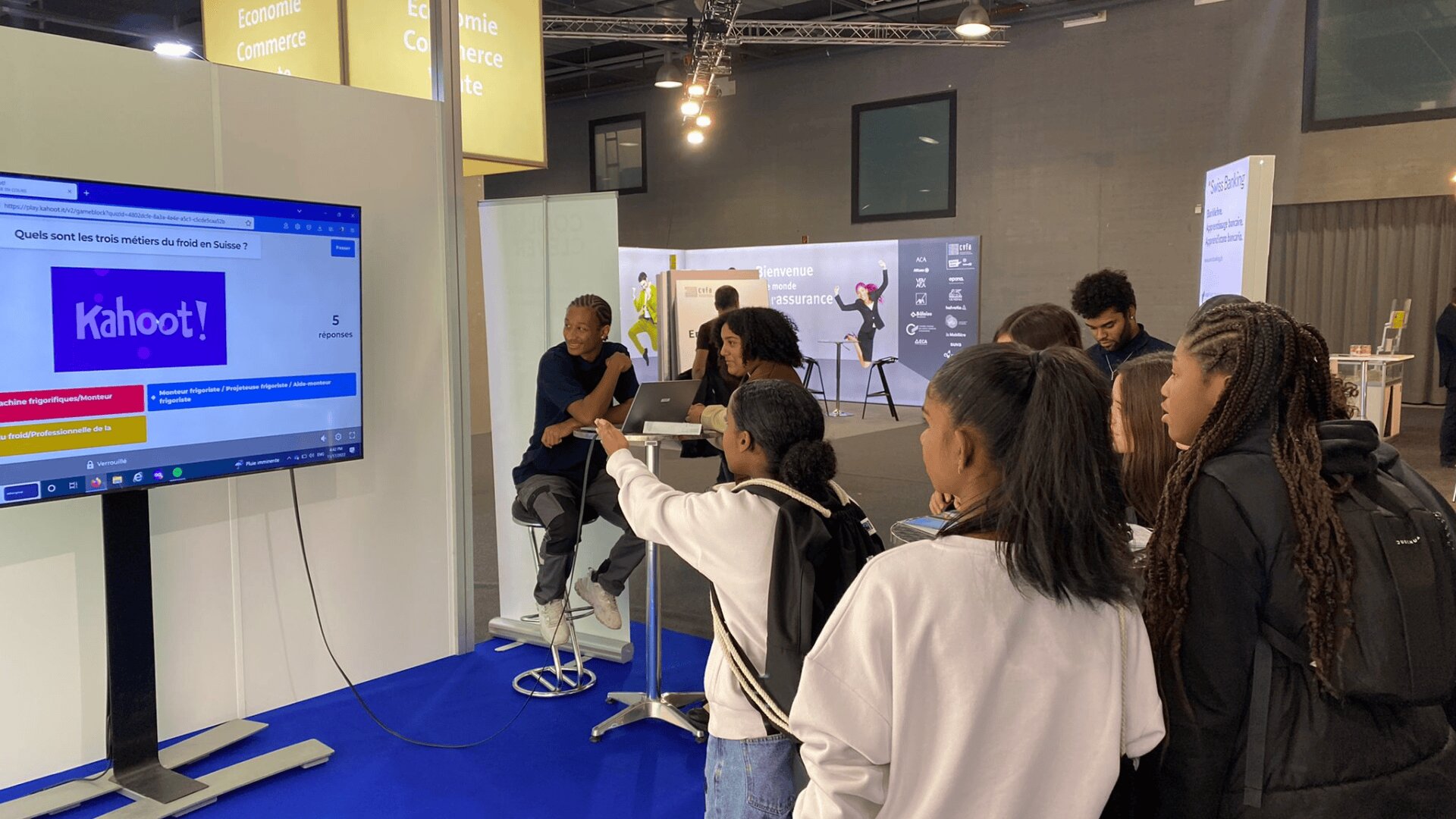 Un groupe d'étudiants se tient devant un écran affichant un quiz Kahoot !, certains levant la main pour participer, tandis que deux présentateurs sont assis à une table haute avec des ordinateurs portables dans un espace d'exposition moderne.